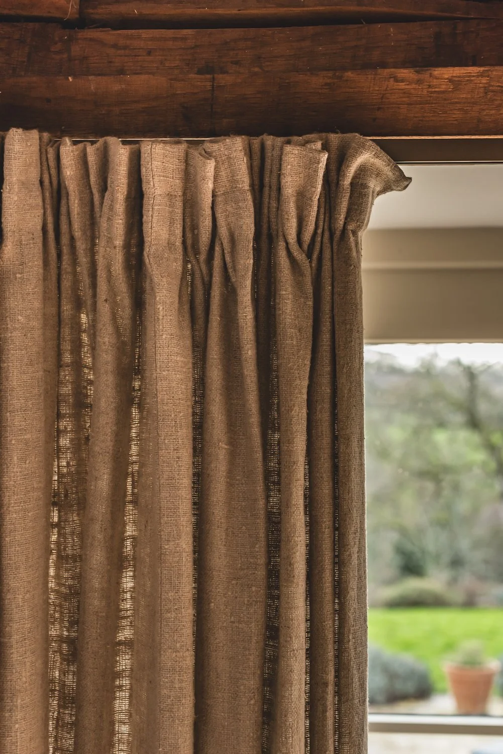 Brown textured luxury pleated curtains hanging in front of a window with a view of trees and a potted plant outside.