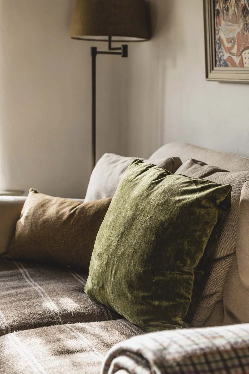Close-up of a luxury beige sofa with green and brown velvet pillows in a living room with a framed artwork and a standing lamp.