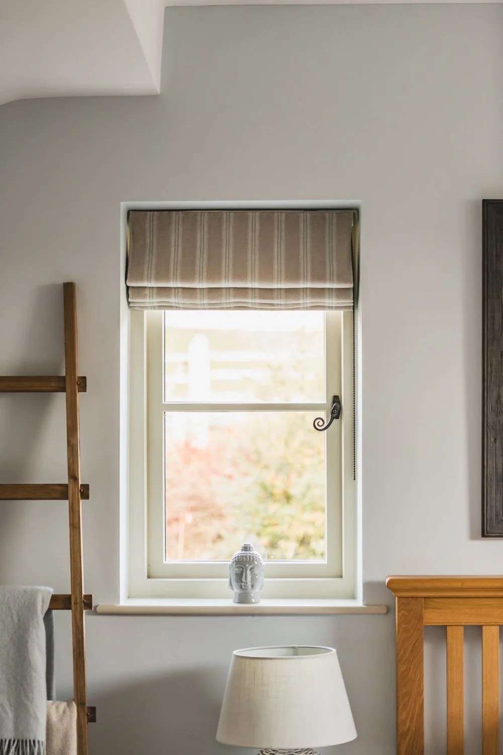 A window with a expertly fitted beige roman blind, a white ceramic Buddha head on the windowsill, and part of a wooden bed frame in a cosy, neutral-toned room.