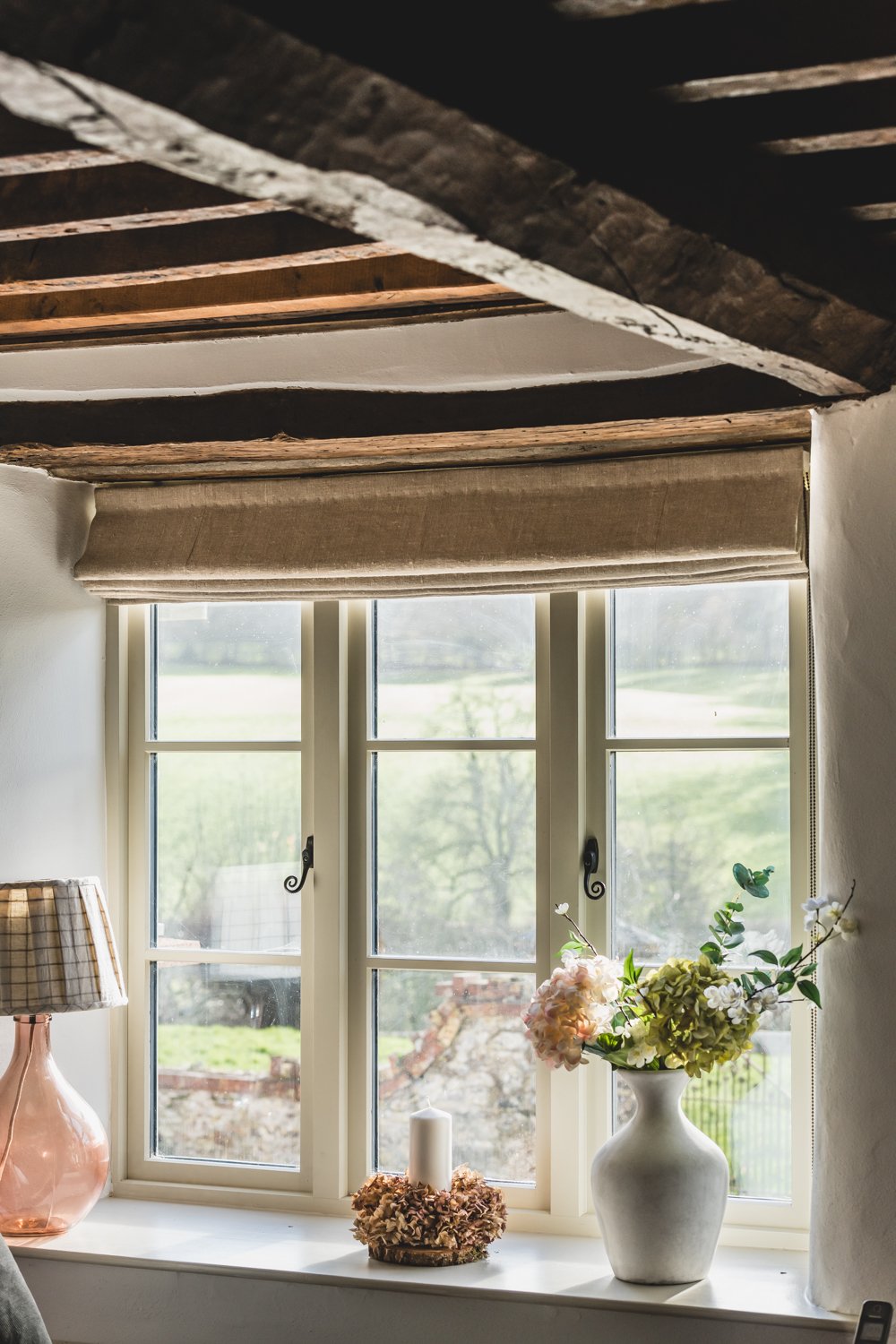 A window with beige blinds above a white windowsill, with a white vase holding flowers, a white candle, a pink glass lamp shade, rustic beams above and a view of greenery outside.