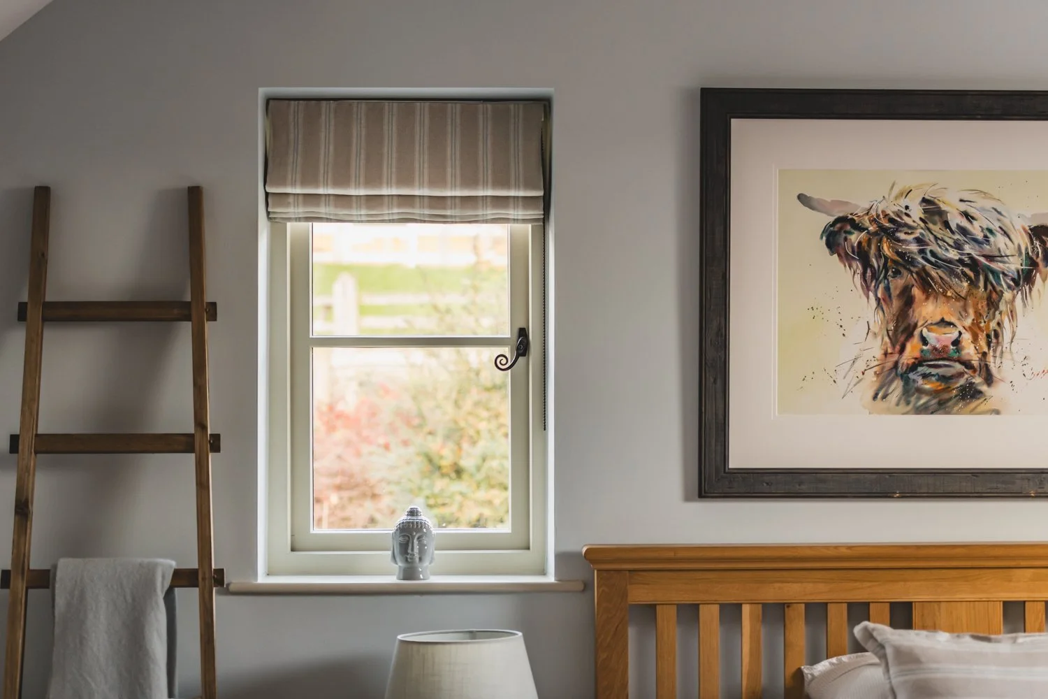 A bedroom wall showing a fitted roman blind in the window, a framed painting of a Highland cow, a wooden ladder with a towel hanging, and a lamp on a bedside table.