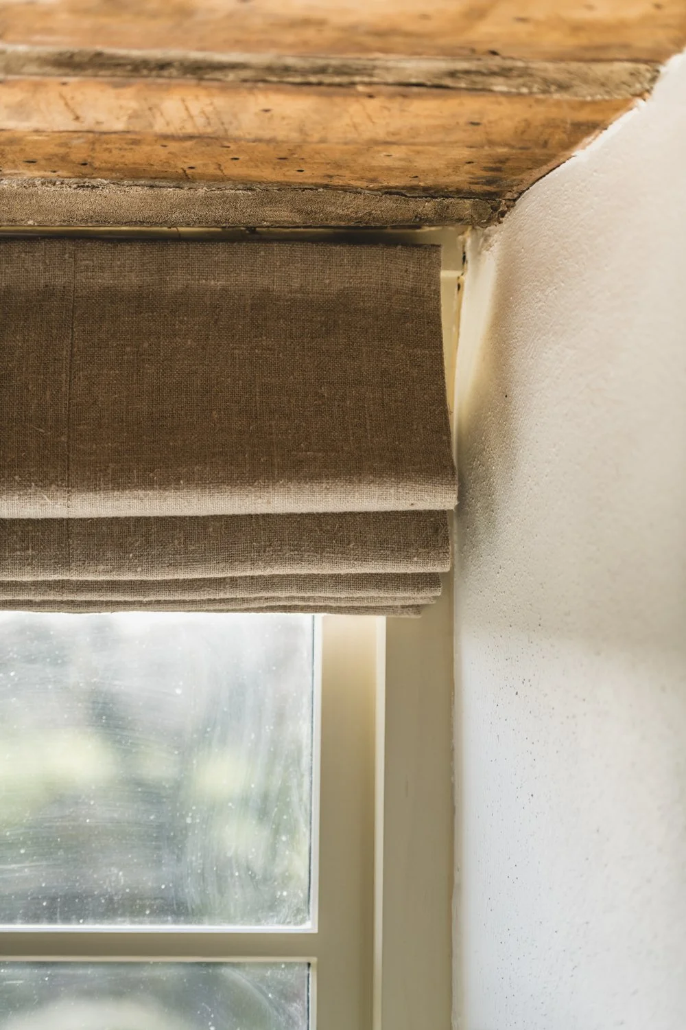 Close-up of a window with a perfectly fitted beige roman blind and a wooden window sill, adjacent to a white wall. Rustic beams and countryside scene.