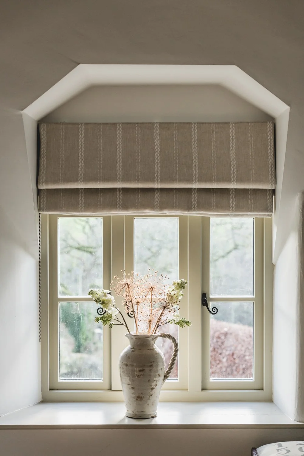 A window with three panes, an expertly fitted beige roman blind, and a white vase with flowers on the windowsill.