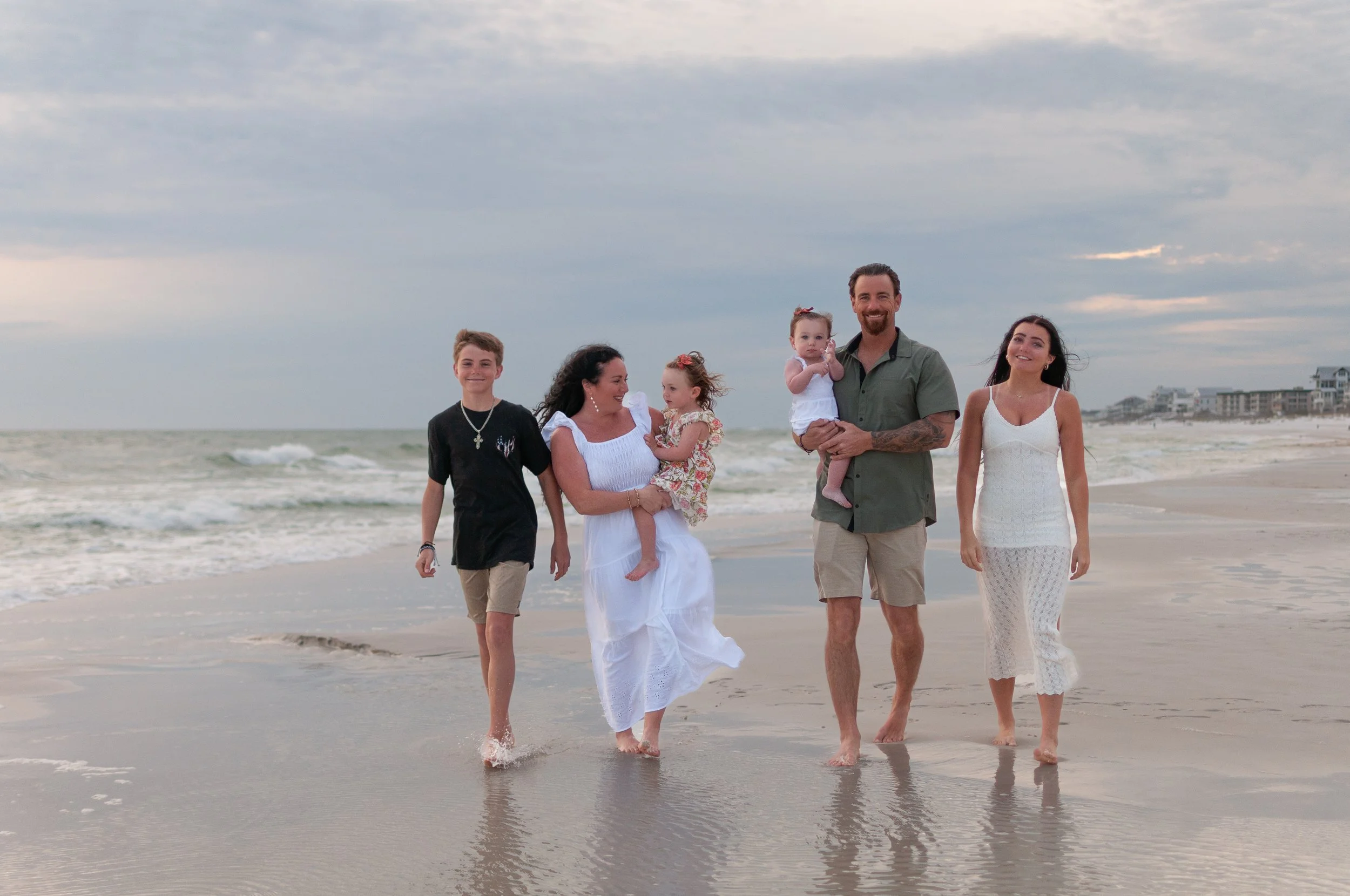 A family of six walking barefoot on the beach, with ocean waves and cloudy sky in the background, smiling and enjoying their time together.