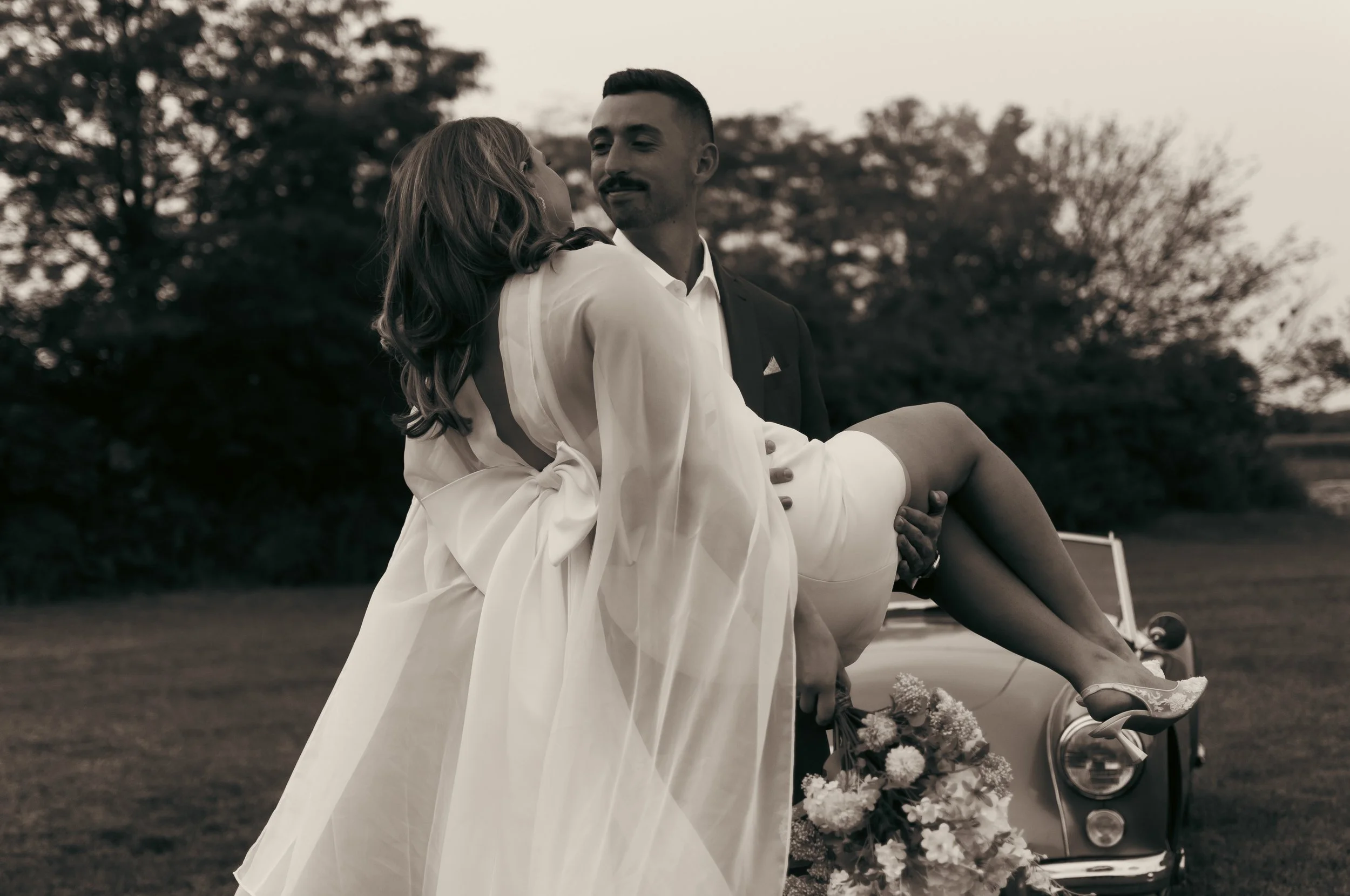 A man in a suit carrying a woman in a wedding dress outdoors with trees in the background, black and white photograph.