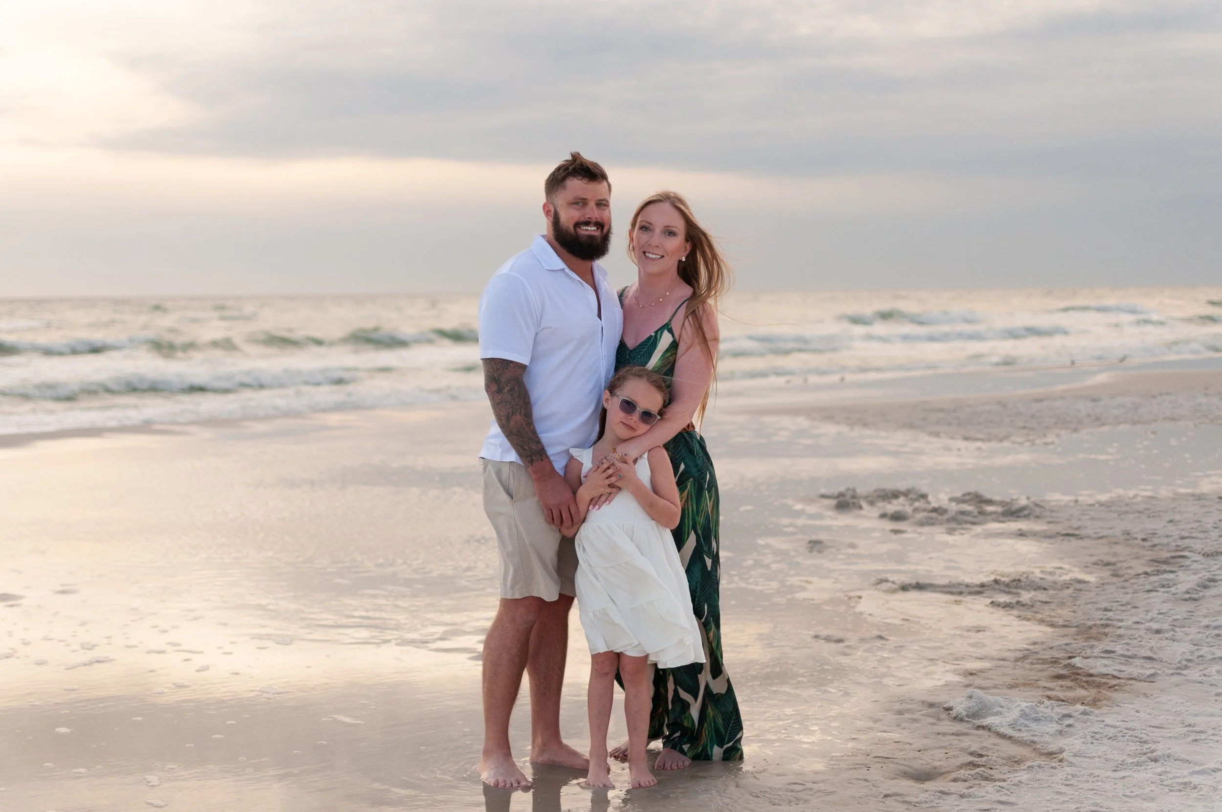 A family of three standing on a beach at sunset: a bearded man in a white shirt and beige shorts, a woman in a long green dress, and a young girl in a white dress with sunglasses.