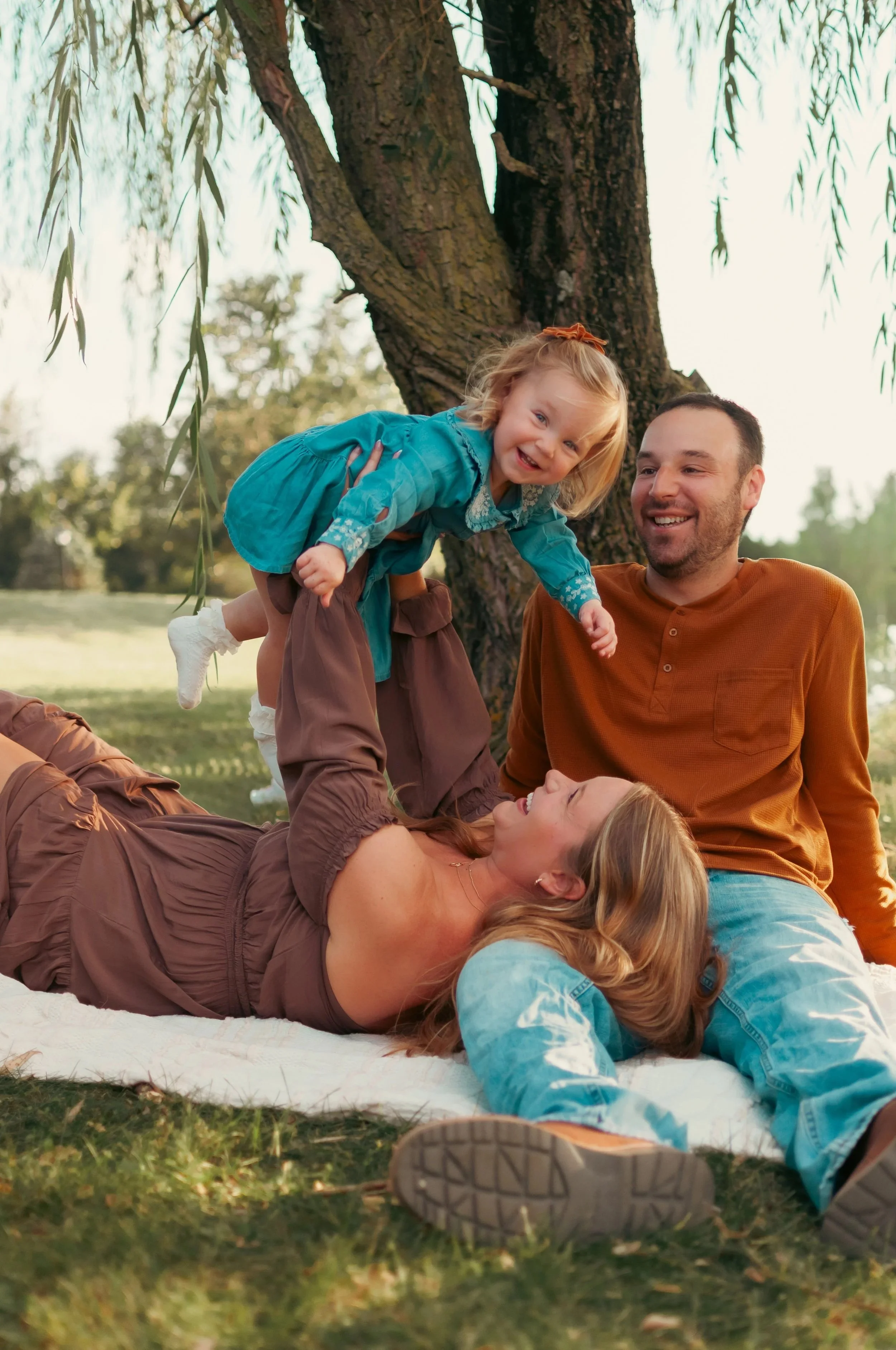 Family of four outdoors playing near a large tree, with a woman lying on the ground, a man sitting nearby, and two young girls, one on the ground and one being held in the air.