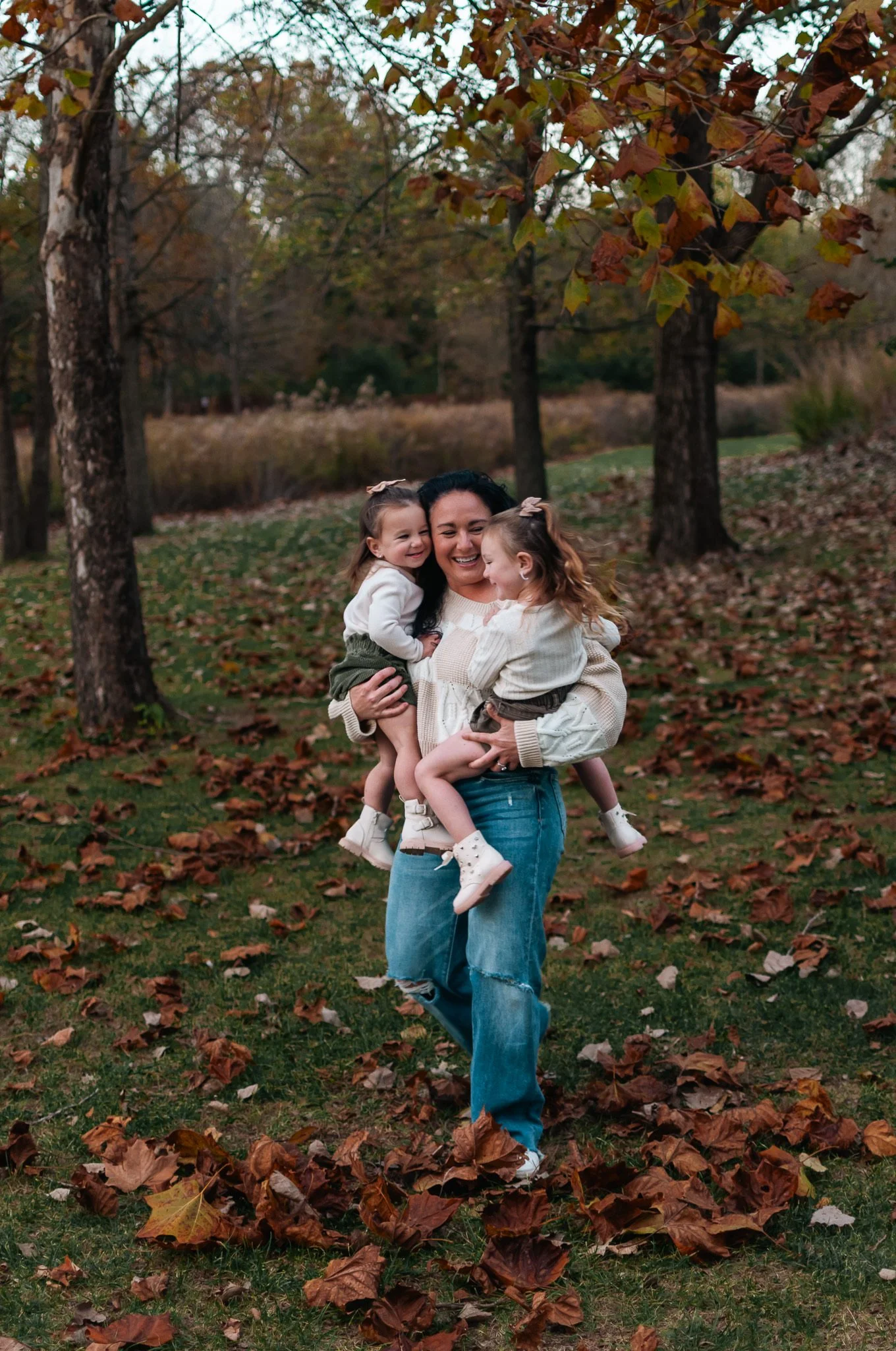 A smiling woman holding two young girls, all wearing cozy sweaters, in an autumn park with fallen leaves on the ground and trees in the background.