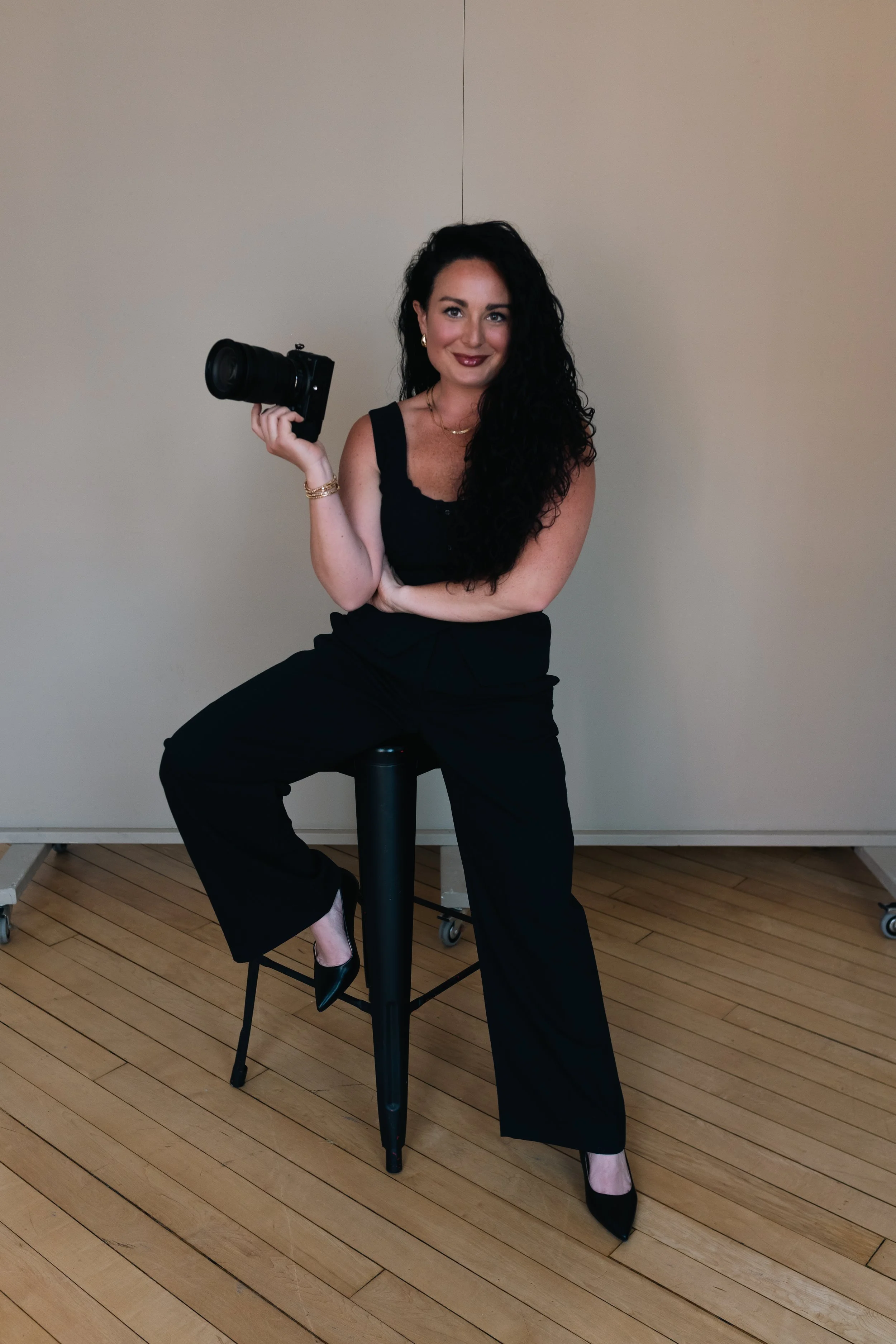 A woman with long black curly hair, dressed in black, sitting on a stool, holding a camera in her right hand, smiling, with a plain light-colored background.