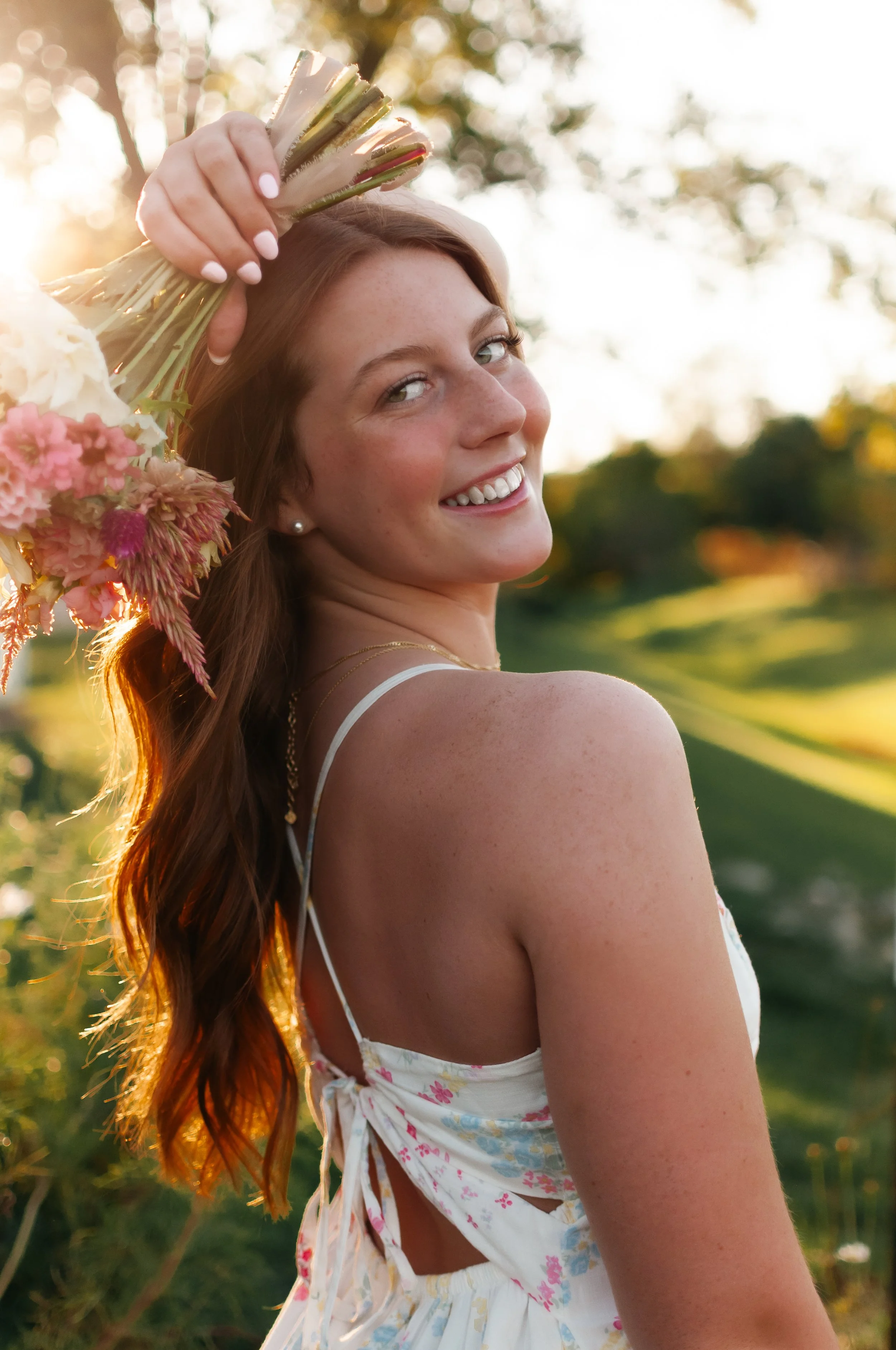 A young woman with long red hair, smiling, holds a bouquet of flowers on her head in a field during sunset.