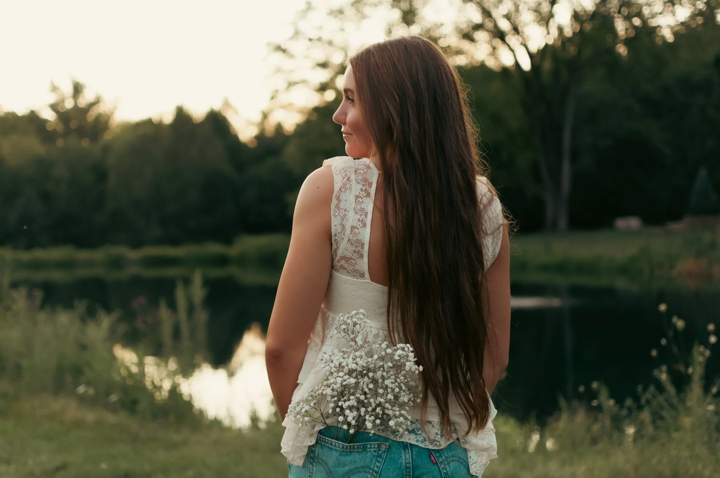 A woman with long brown hair in a cream-colored lace top and blue jeans near a lake at sunset, holding a small bouquet of white baby's breath flowers.
