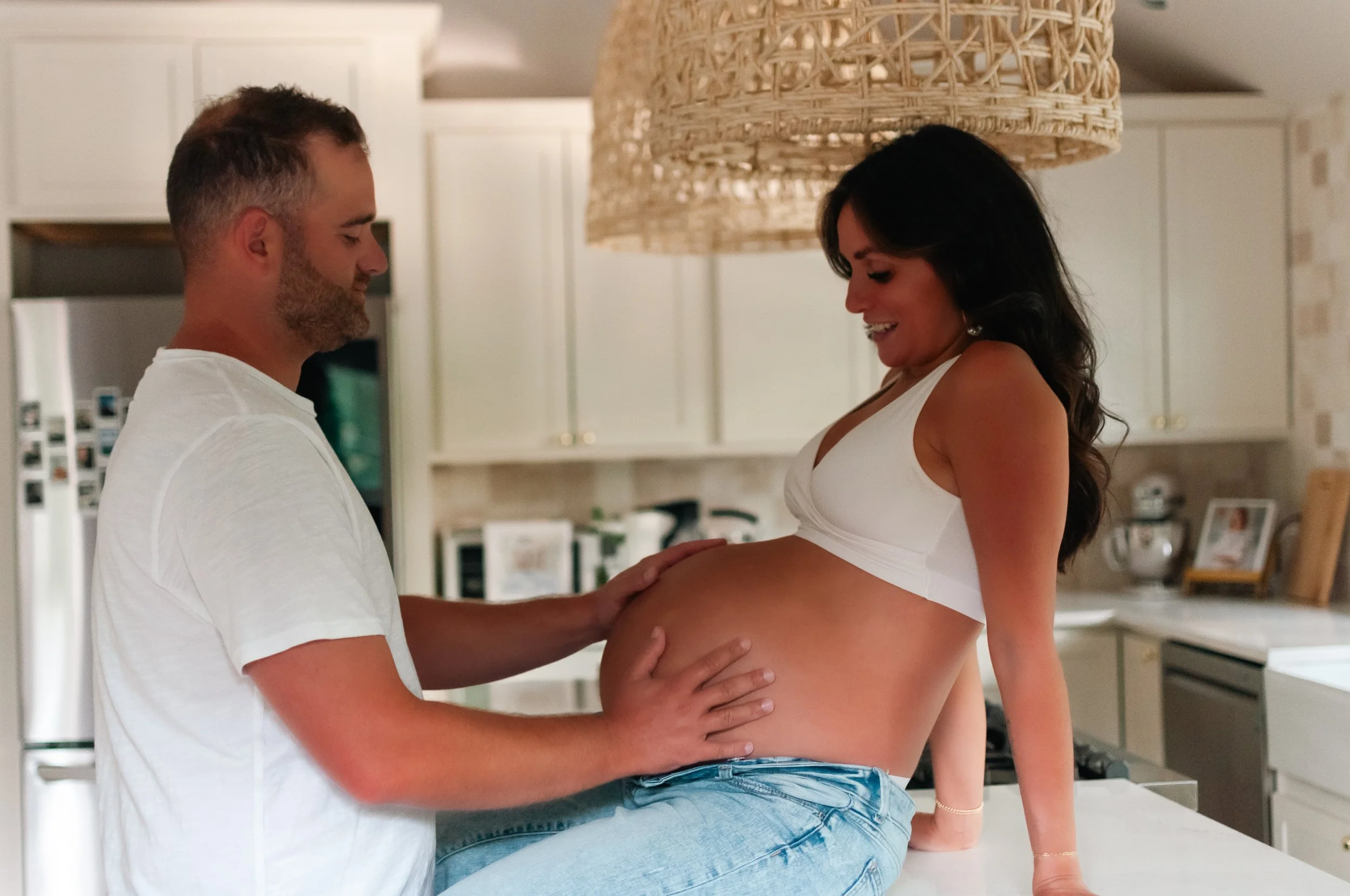 A pregnant woman sitting on a kitchen counter smiling at a man who is touching her belly.