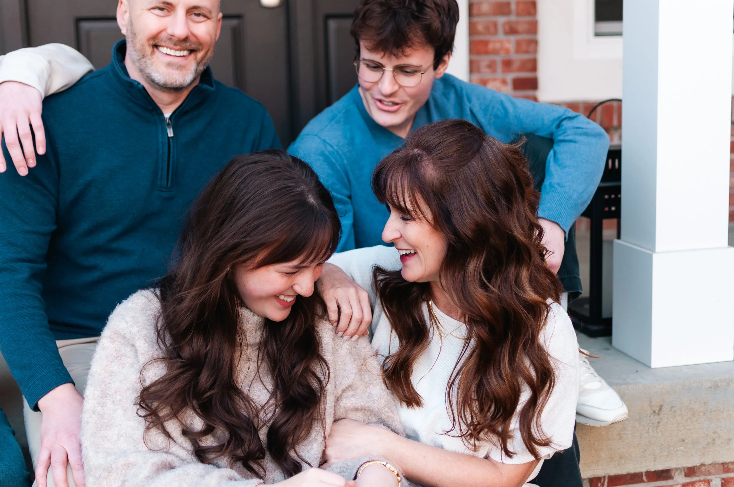 A family of five gathering outdoors on a brick porch, smiling and laughing, with two young women in the front and two men standing behind them.