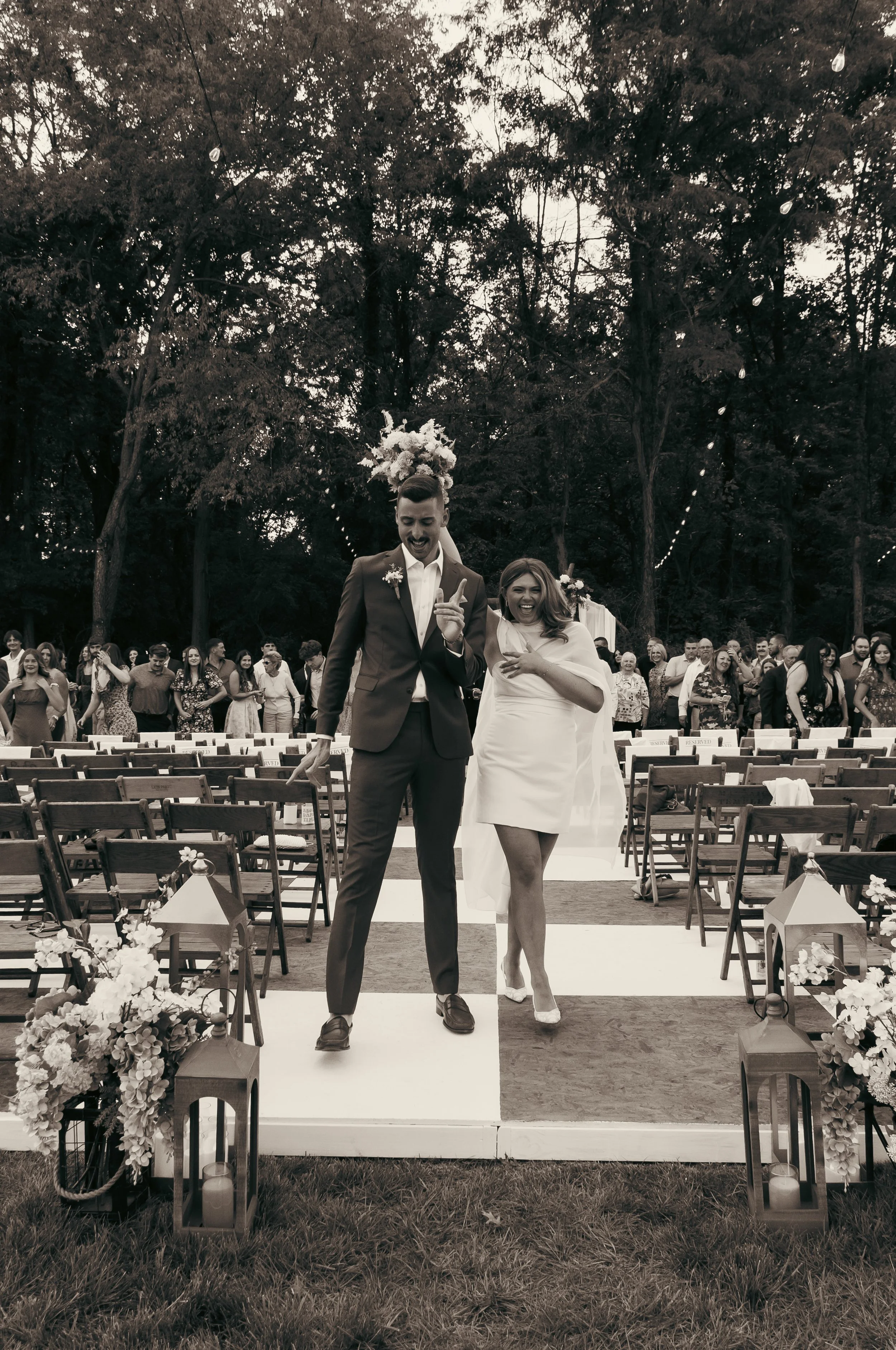 A couple smiling and walking down the aisle at an outdoor wedding ceremony. The groom is dressed in a suit and the bride in a white dress. Guests are standing behind them, and there are chairs, flowers, and lanterns along the aisle.