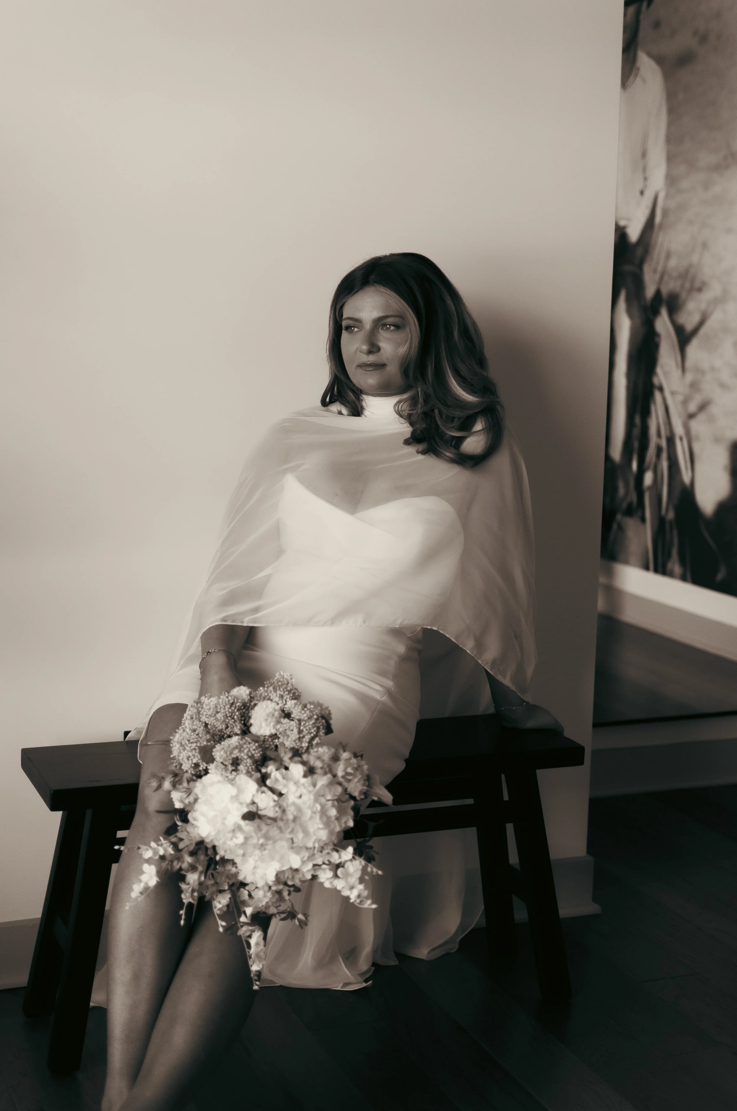 A woman in a wedding dress sitting on a bench, holding a bouquet of flowers, in black and white.