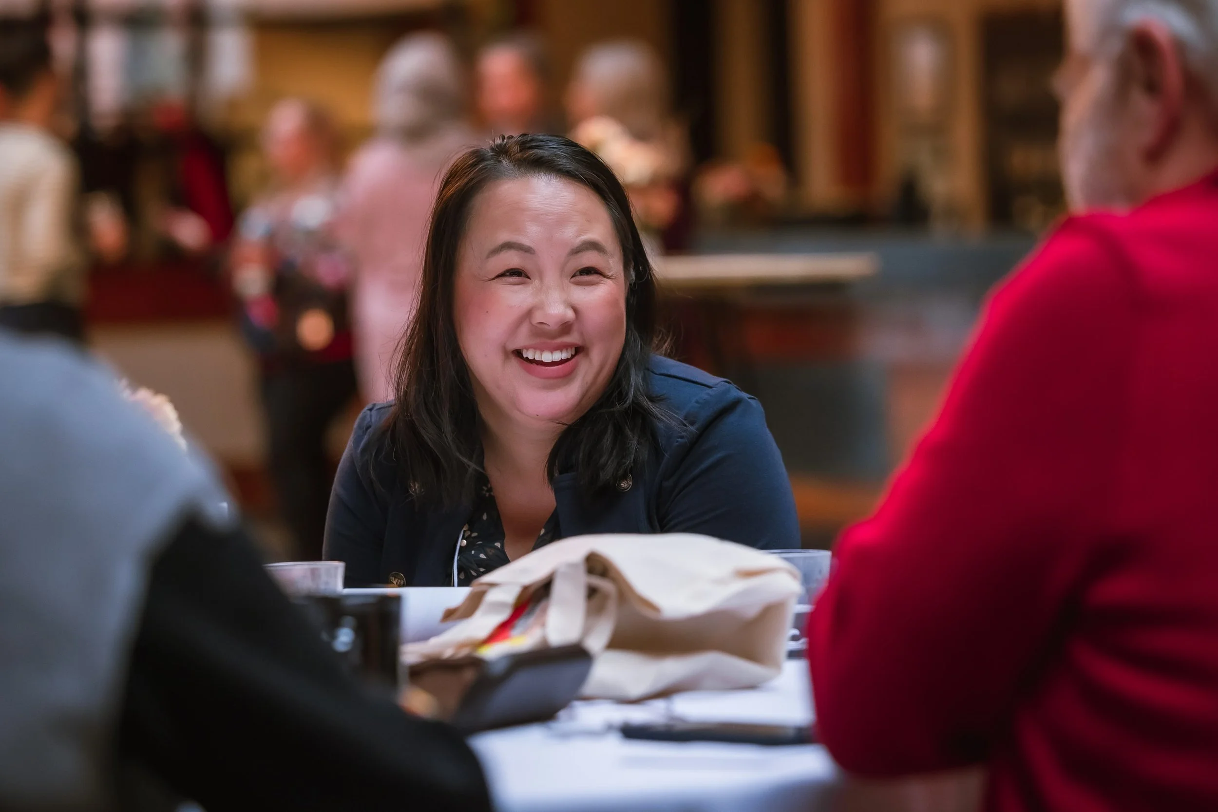 A woman with dark hair smiling while talking with two other people at a table in a lively restaurant or cafe.