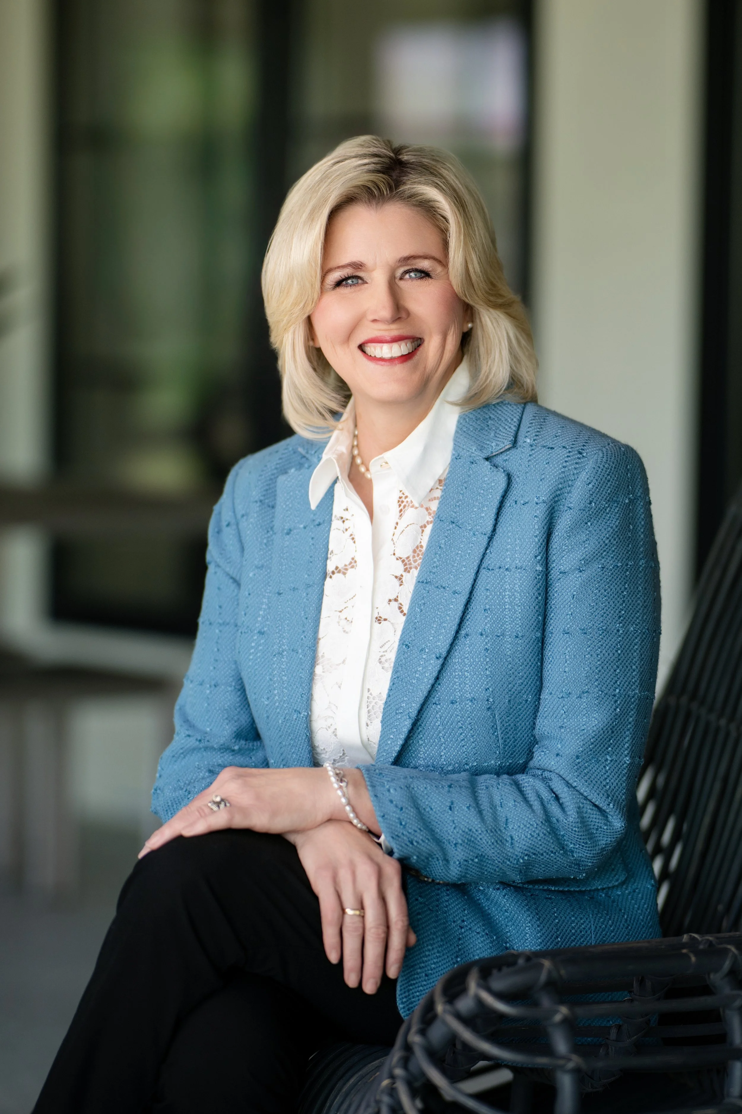 A smiling blonde woman wearing a blue blazer, white blouse, and pearls, sitting outdoors on a black wicker chair with a blurred background.