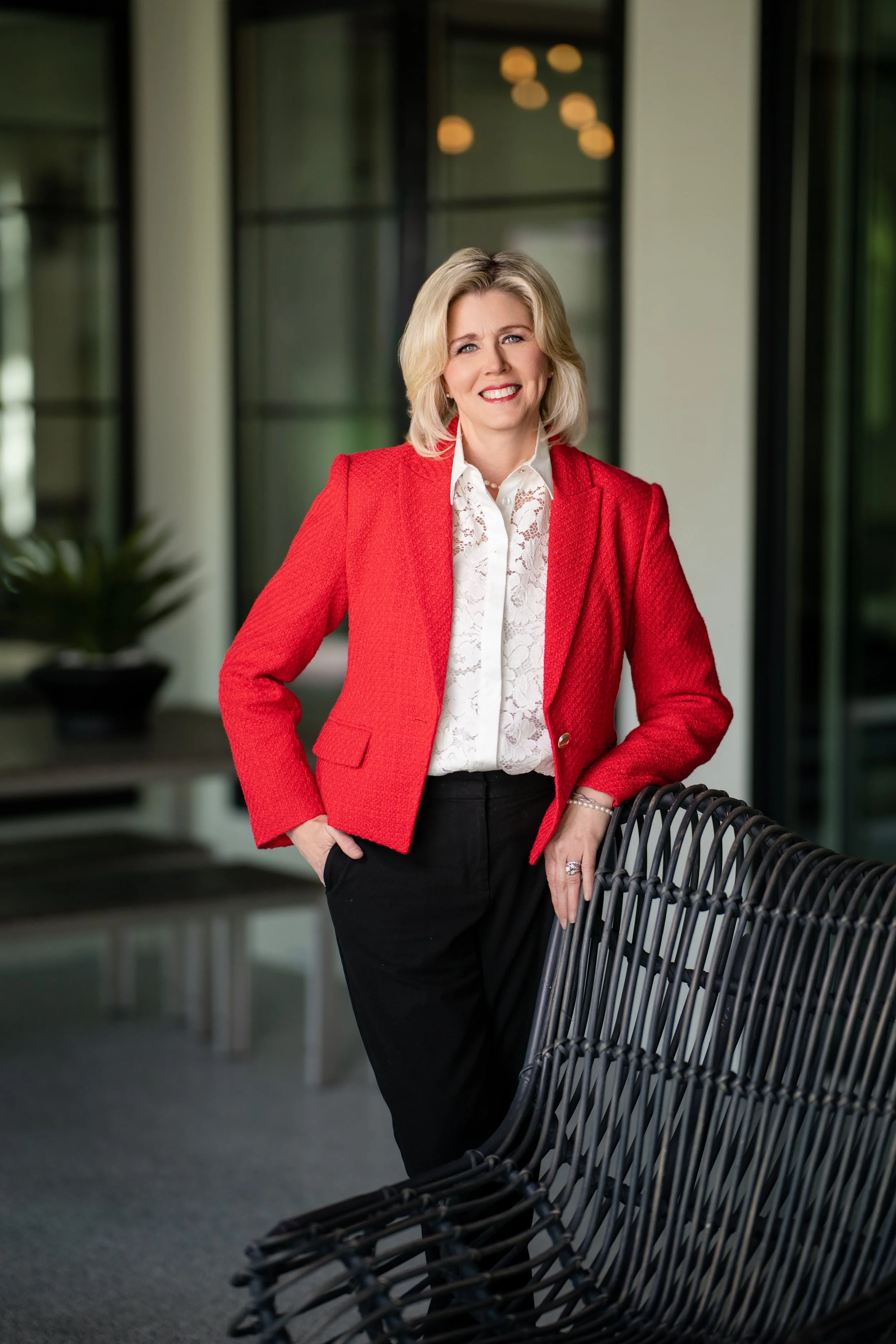 A woman with blonde hair, wearing a red blazer over a white lace blouse, black pants, and a pearl necklace, standing beside black outdoor chairs in a modern glass-walled building.