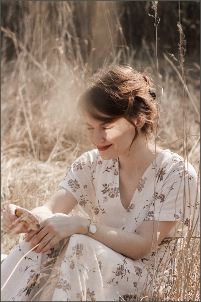A woman with brown hair and a floral dress sits in a field of dry grass, smiling while looking at her phone.