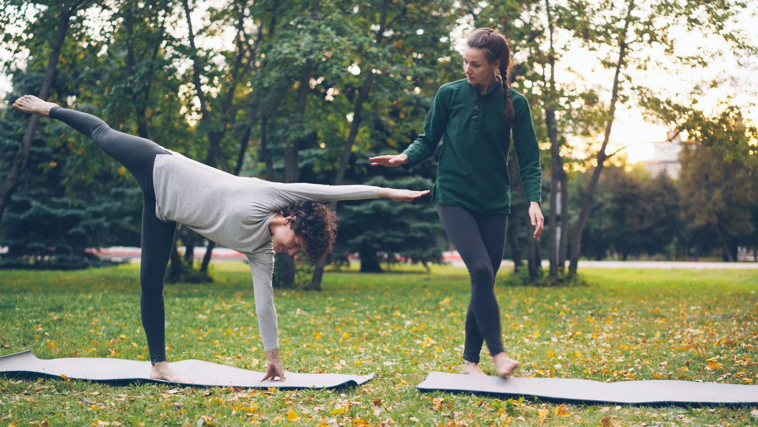 A woman practicing yoga outdoors on a mat in a park during autumn, assisting a young girl in balancing in a yoga pose.
