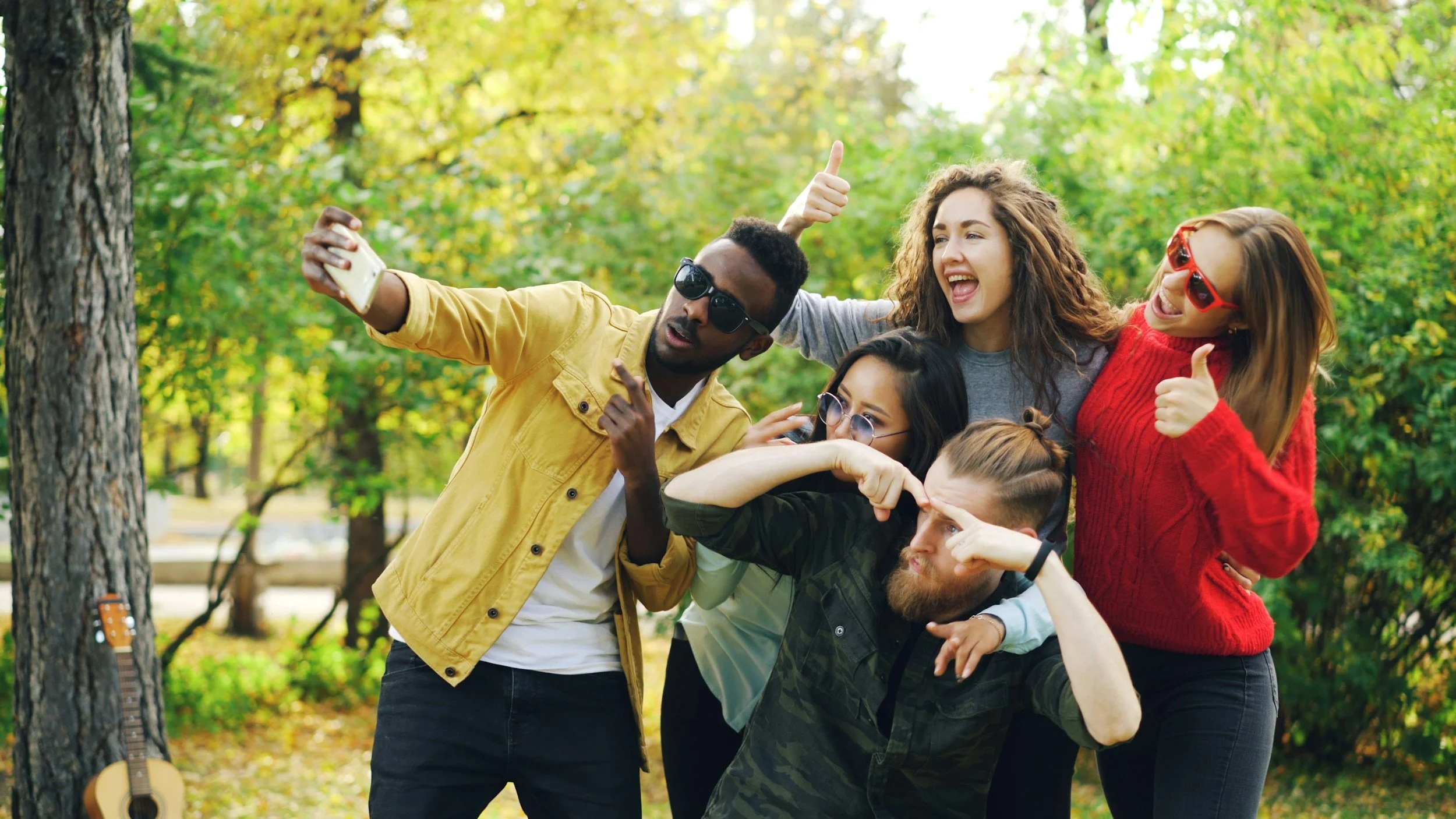 Group of five young friends taking a selfie together in a park during daytime, all smiling and making various gestures, with trees and greenery in the background.