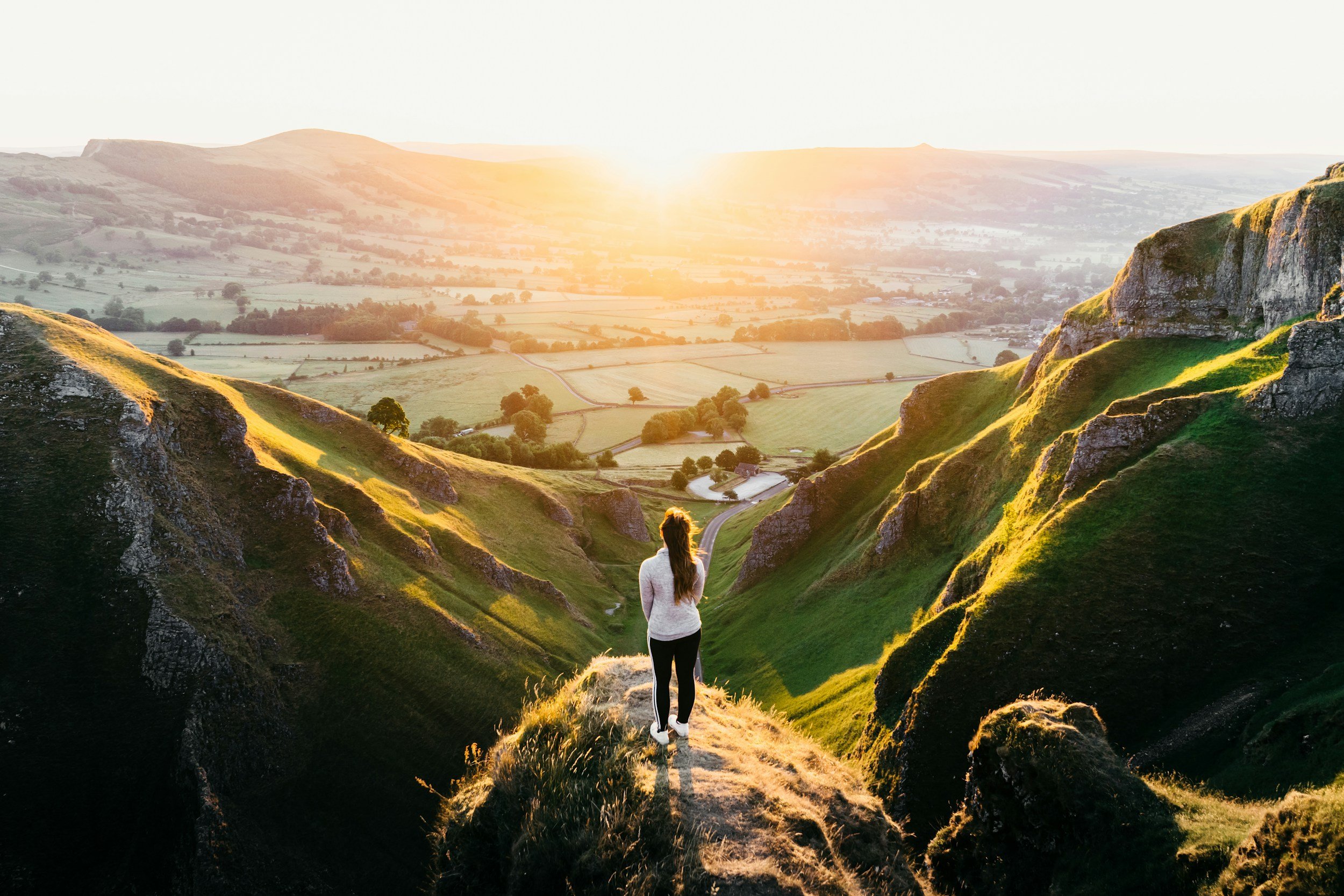 A woman walking through a scenic valley with green hills and rocks during sunset.