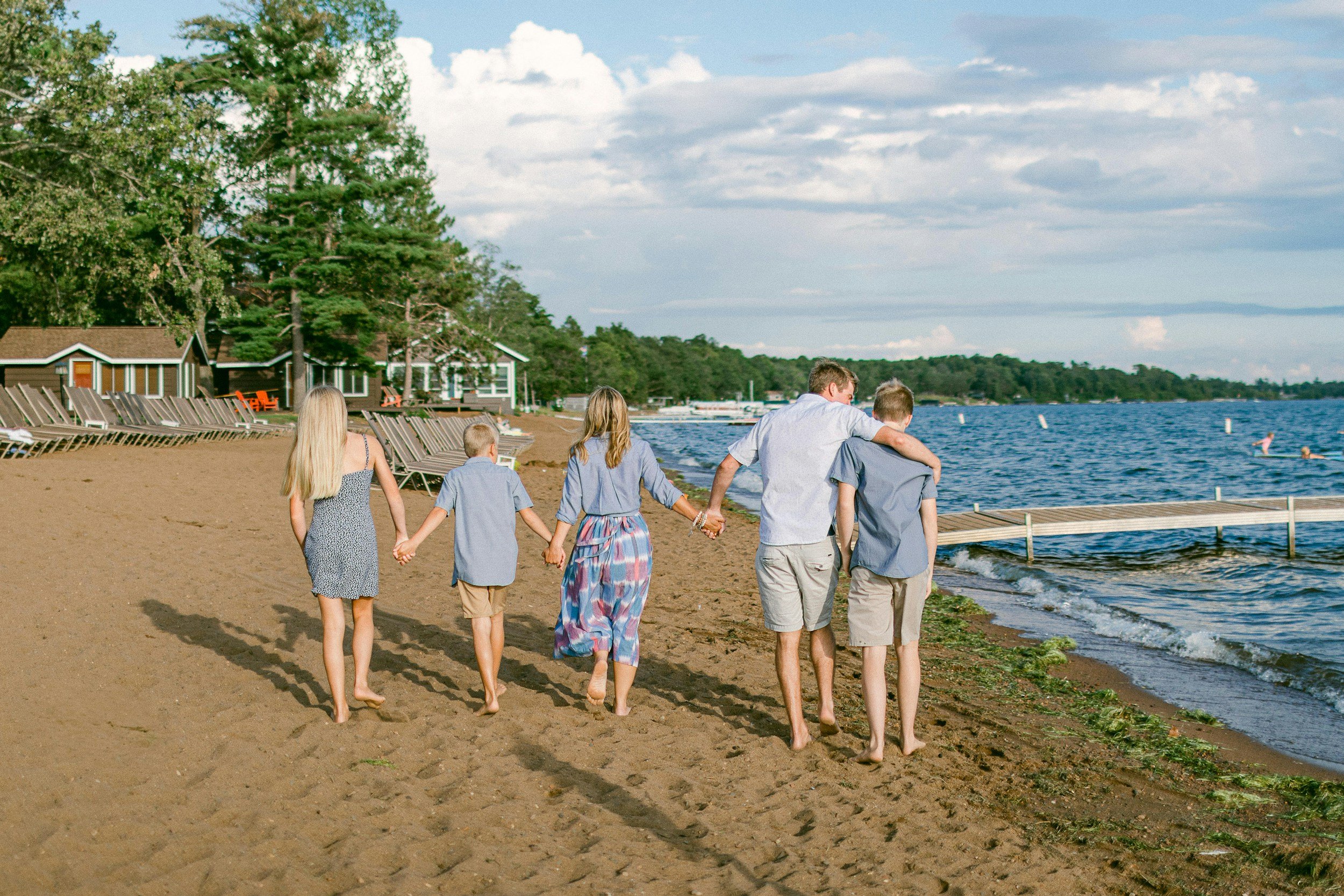 A family of six with three children and two adults walking barefoot along a sandy beach holding hands, with houses, trees, and a body of water in the background on a partly cloudy day.