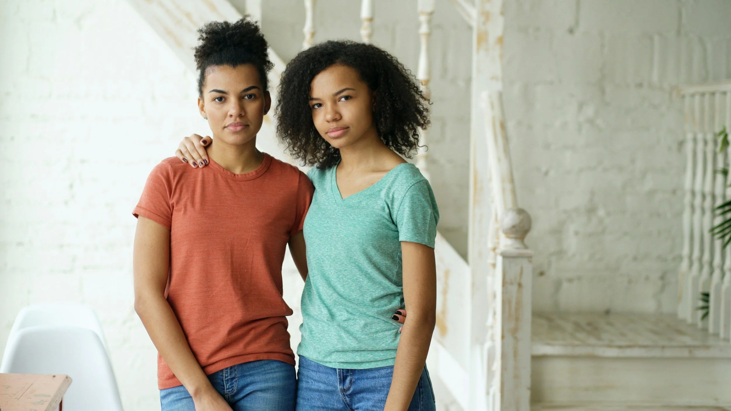 Two young women standing close together indoors, with one woman gently resting her hand on the other's shoulder. The woman on the left is wearing a reddish-brown T-shirt, and the woman on the right is wearing a light green T-shirt. They are standing in front of a white wall with a staircase in the background.