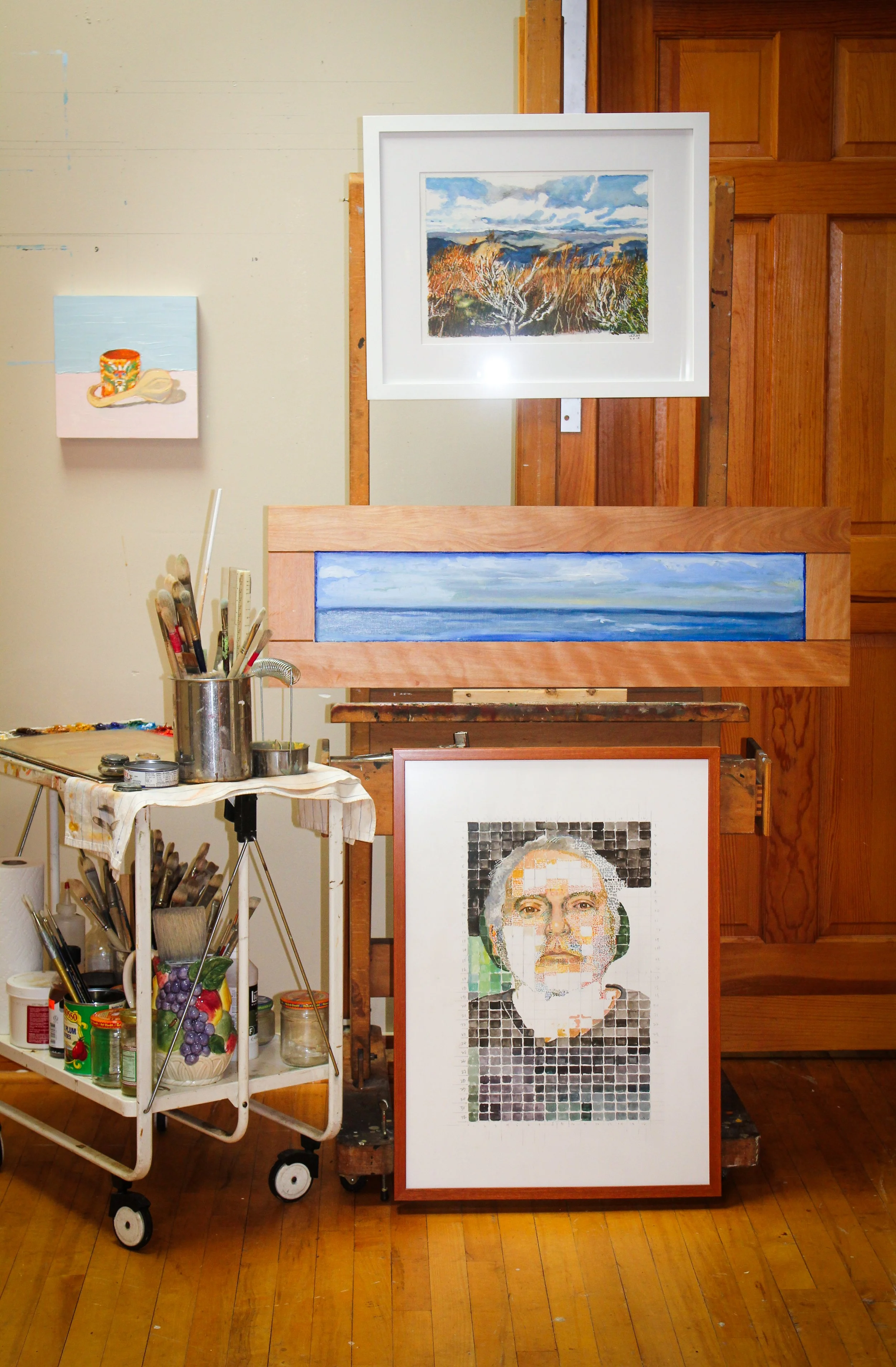 Dean Vallas' artist's studio with paintings displayed on the wall and a portrait being created on an easel. There are brushes and art supplies on a rolling cart. The paintings include a landscape with clouds, trees, and a body of water.