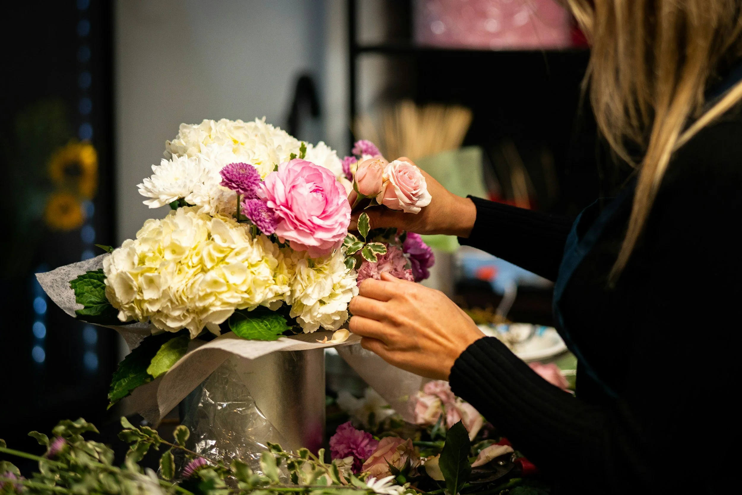 A person arranging a bouquet of white, pink, and purple flowers in a floral shop.