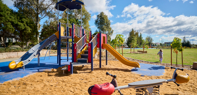 Colorful playground with slides, climbing structures, and a spring rider on a sandy surface in a park with trees and green grass under a partly cloudy sky.