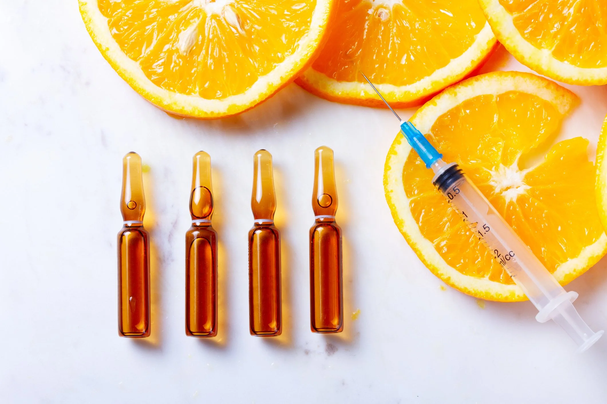 Four glass ampoules filled with an amber-colored liquid, four orange slices, and a syringe with a blue needle cap on a white surface.