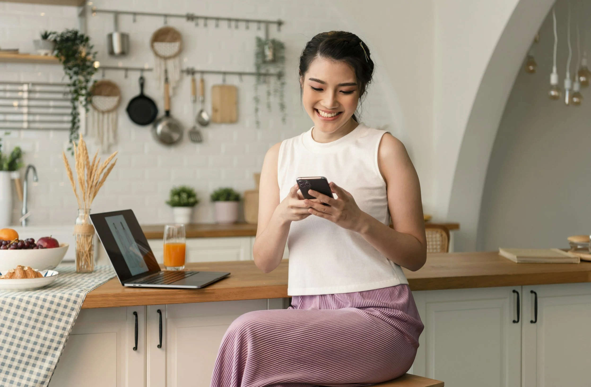 A woman sitting on a kitchen counter using her phone, with a laptop, orange juice, and breakfast items nearby, in a modern kitchen with hanging utensils and potted plants.