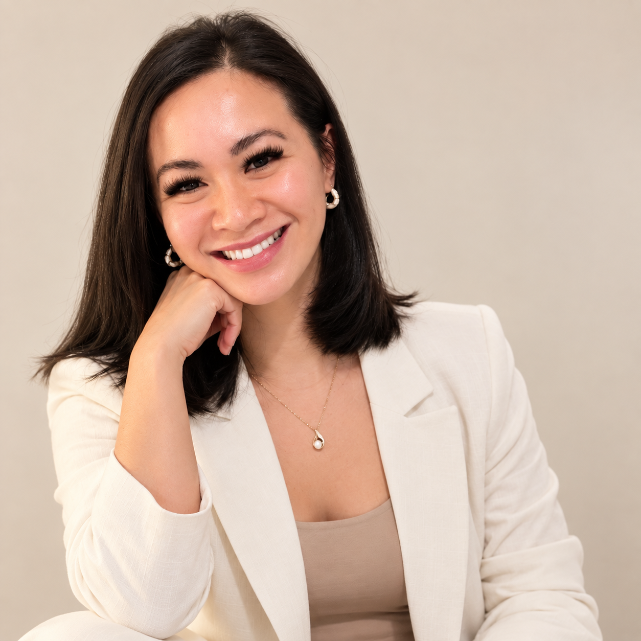 A young woman with shoulder-length dark hair, smiling, wearing a white blazer and beige top, with jewelry including earrings and a necklace, resting her chin on her hand against a plain light background.