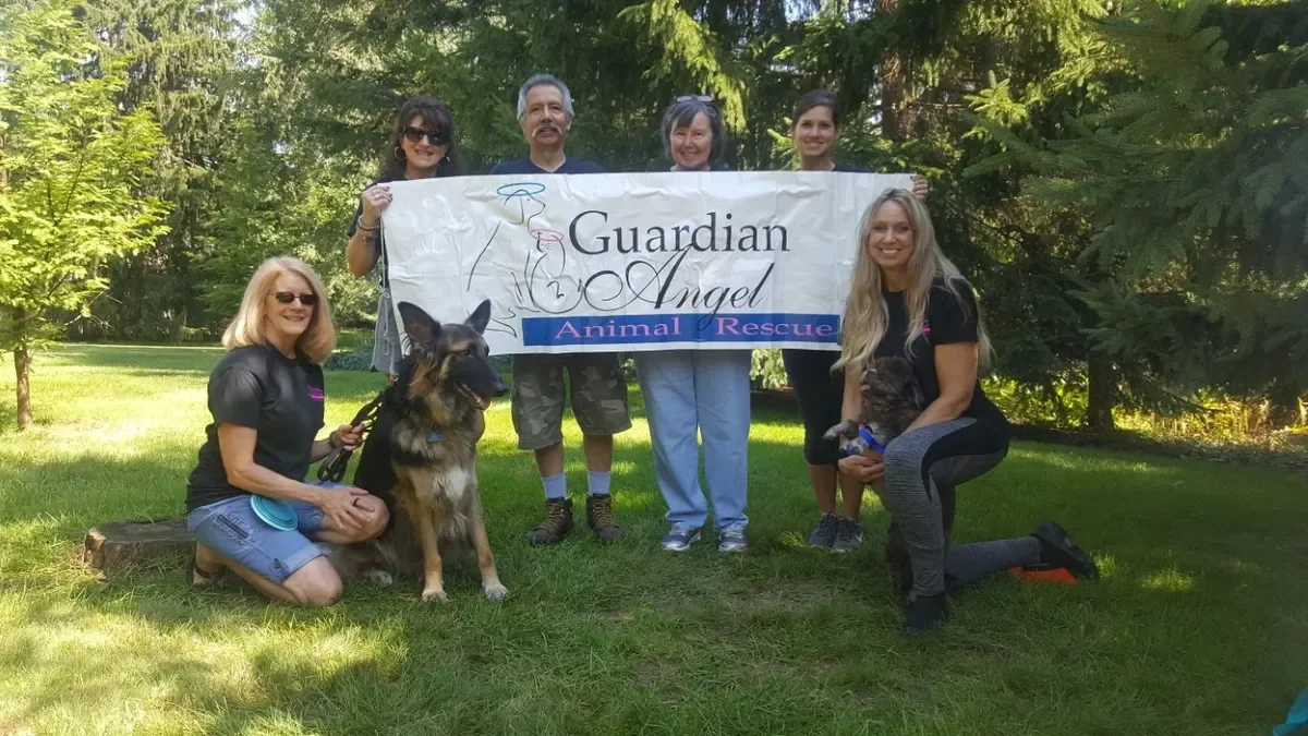 Group of people outdoors holding a banner that reads "Guardian Angel Animal Rescue" with some dogs and smiling.