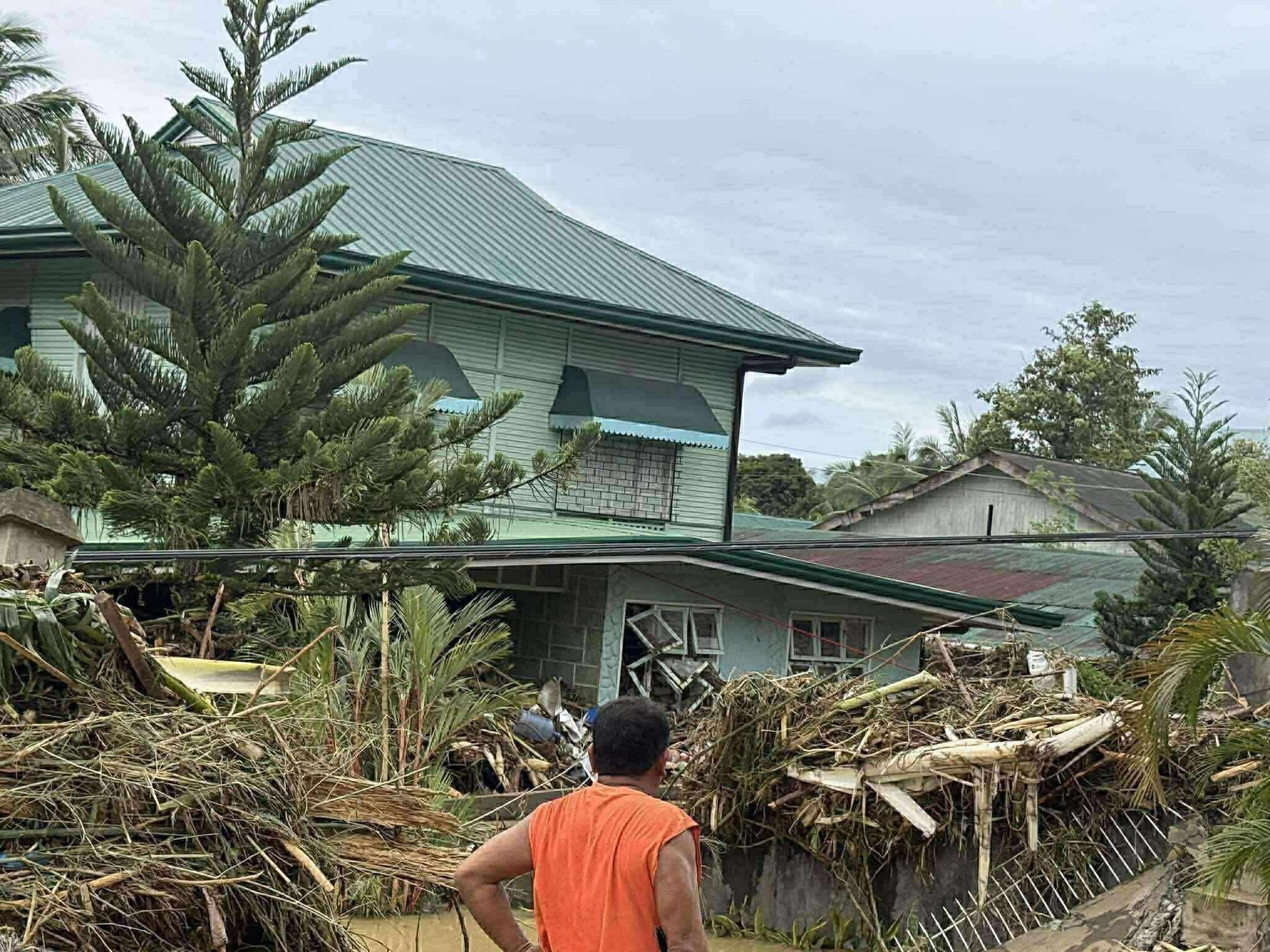 A man in an orange shirt stands in front of a damaged residential area with debris and fallen trees after a storm or hurricane. There are damaged houses and overcast skies in the background.