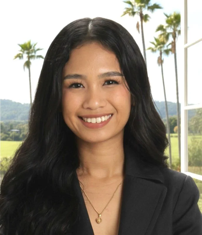 A young woman with long black hair, smiling, wearing a black blazer and a gold necklace, with a background of palm trees and a window overlooking a green landscape.