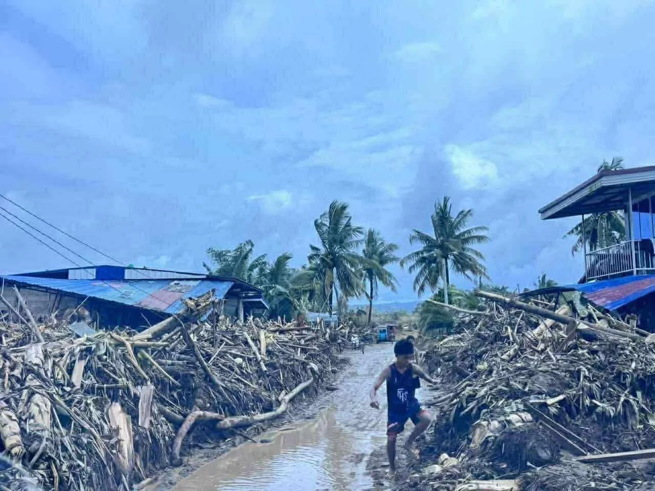 Child playing in muddy street amid destroyed buildings and fallen palm trees after a storm or hurricane.