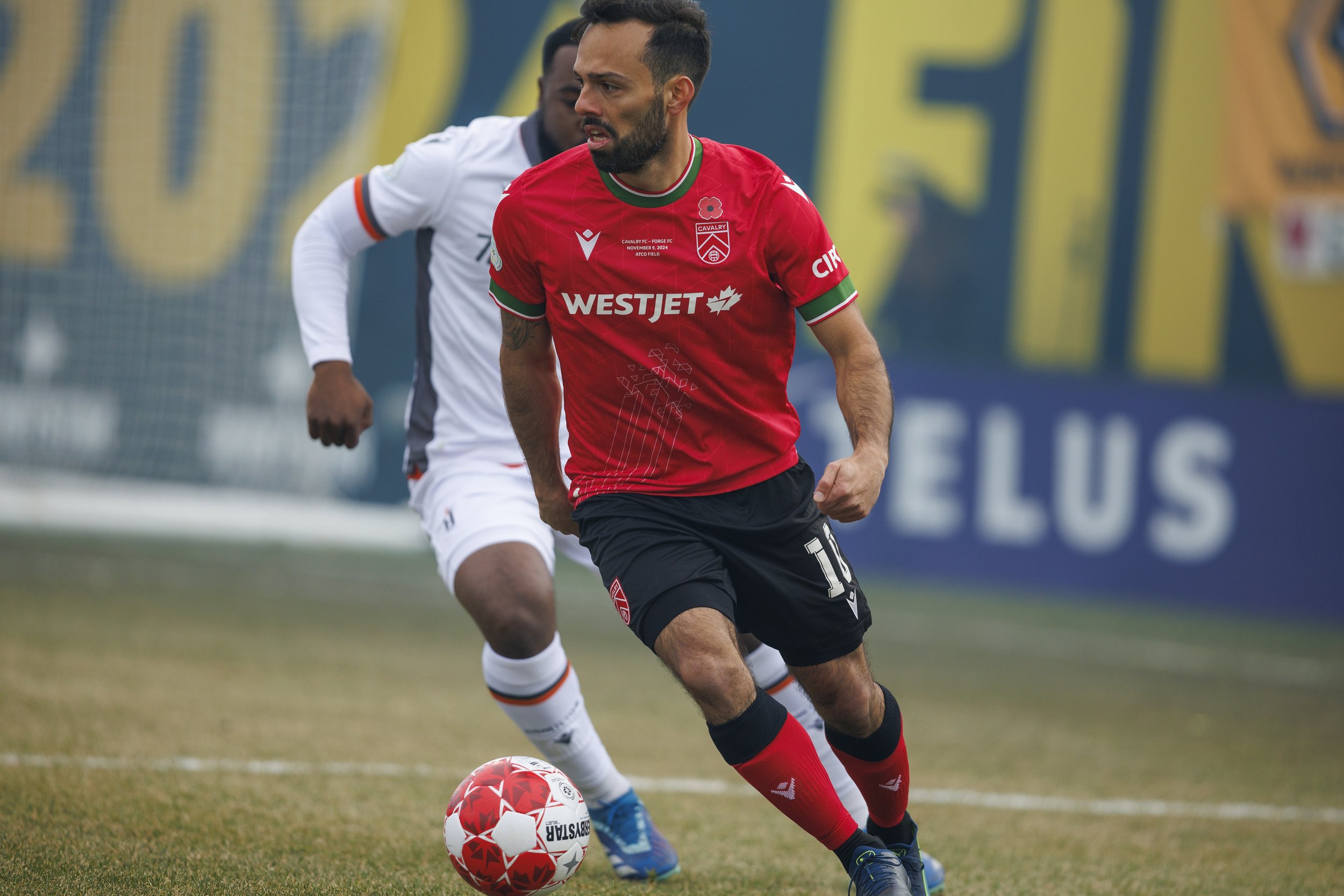 A soccer player in a red jersey and black shorts dribbling a soccer ball during a match, with a player in white in the background.