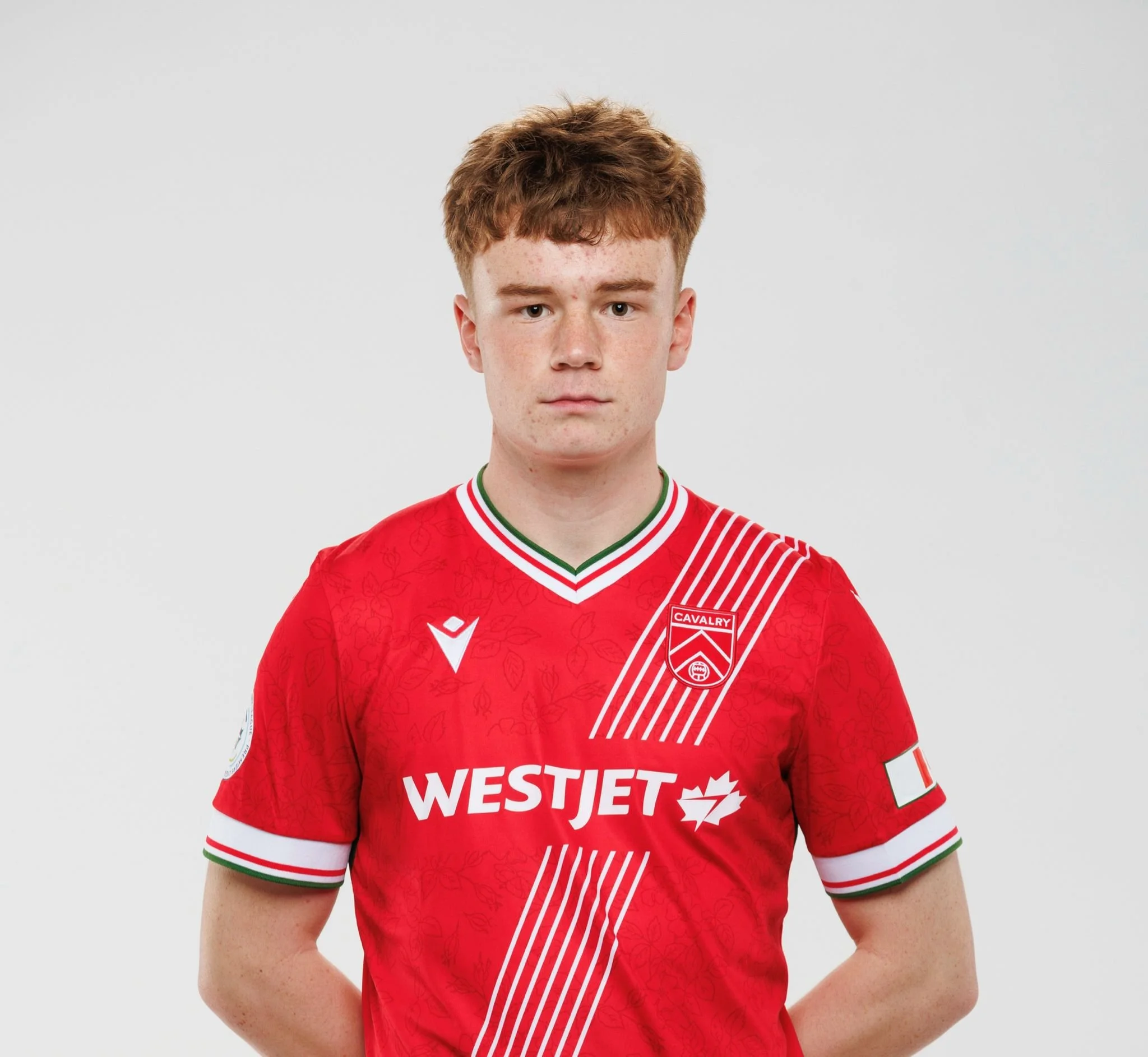 James McGlinchey Cavalry FC soccer player with short red hair wearing a red jersey with white and green accents and white sponsor logo, standing against a plain white background.