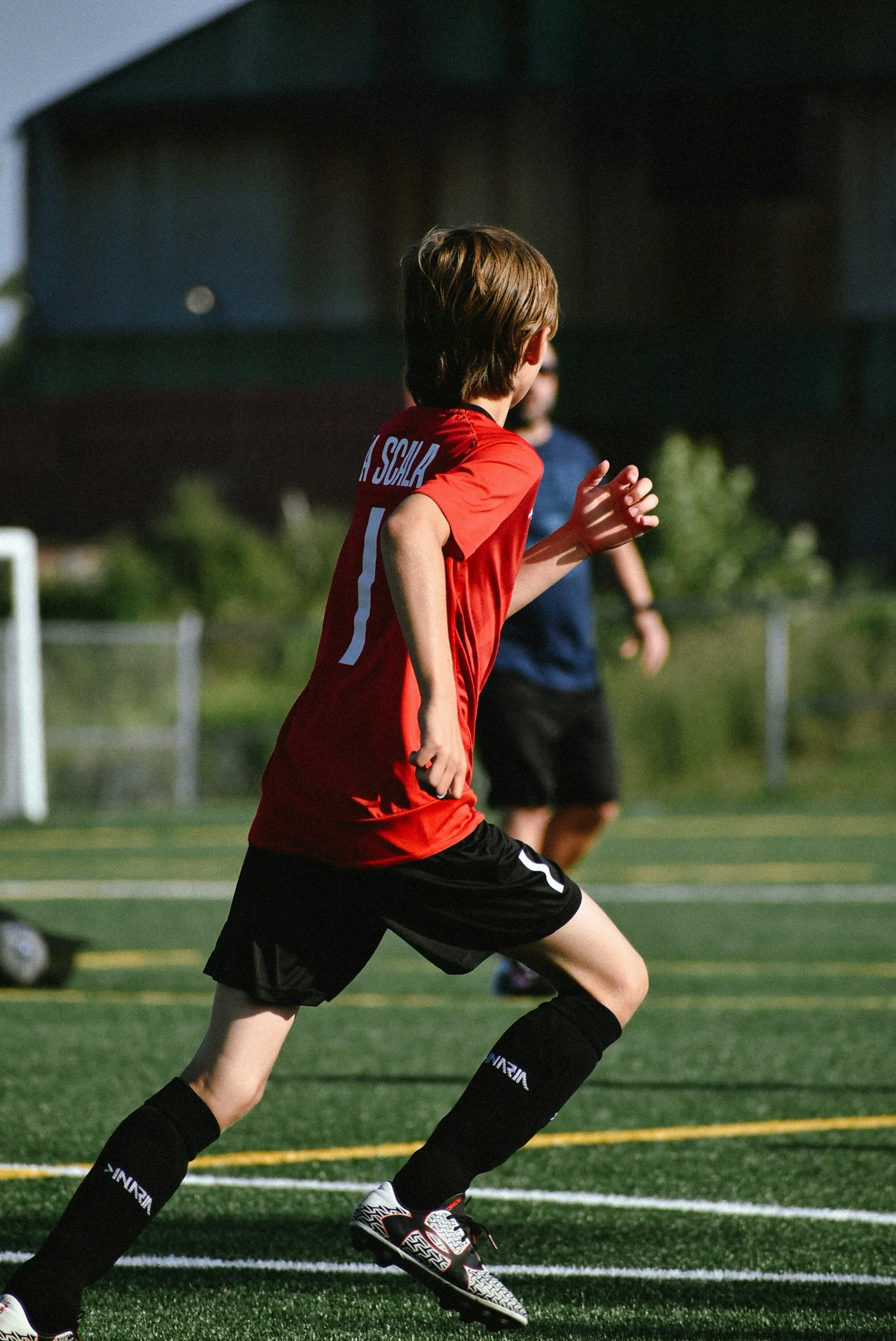 A young boy playing soccer outdoors on a field, wearing a red jersey and black shorts, with other players and a building in the background.