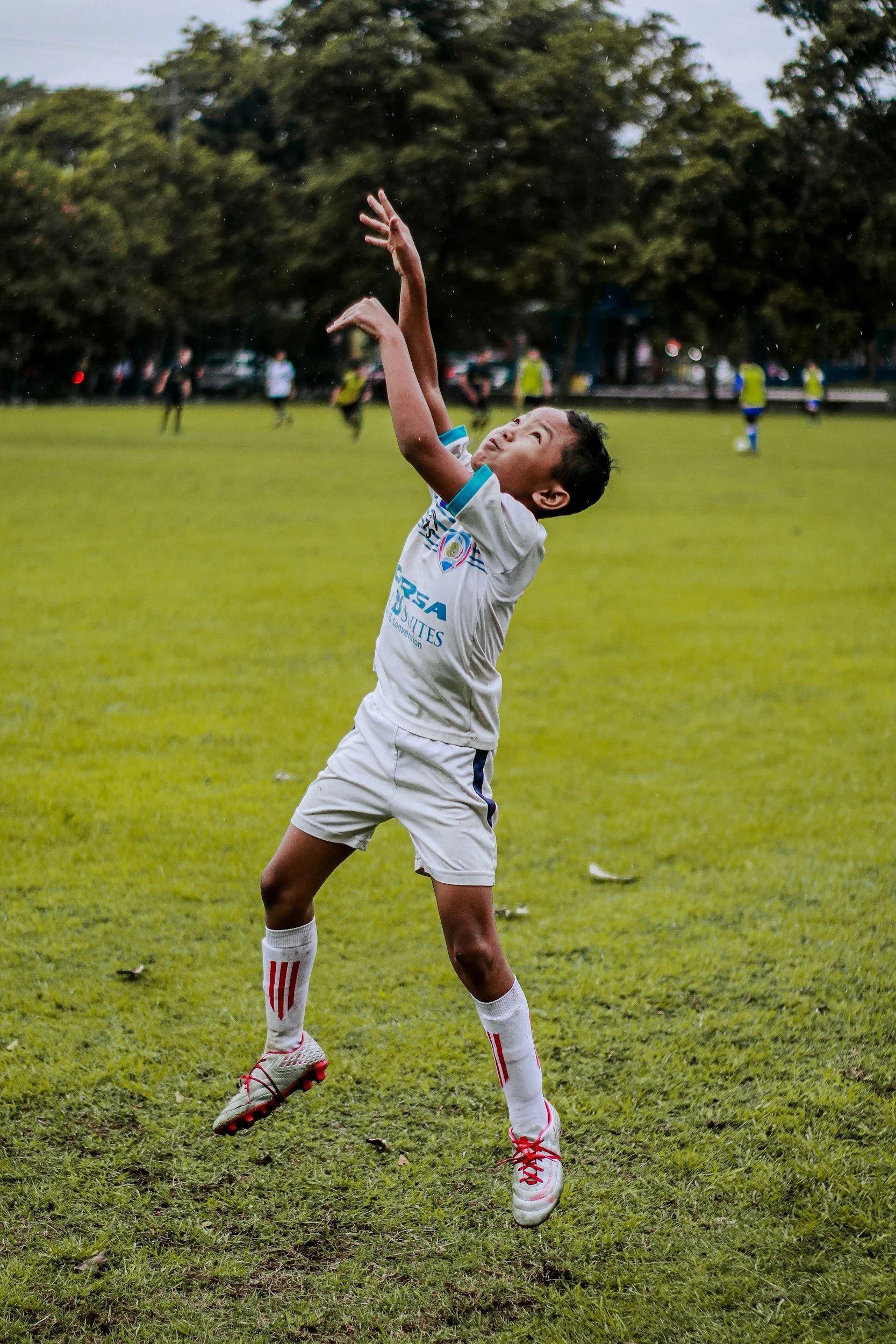 A young boy in a white soccer uniform with red laces jumps to catch a ball on a grassy field during a game, with other players and trees in the background.