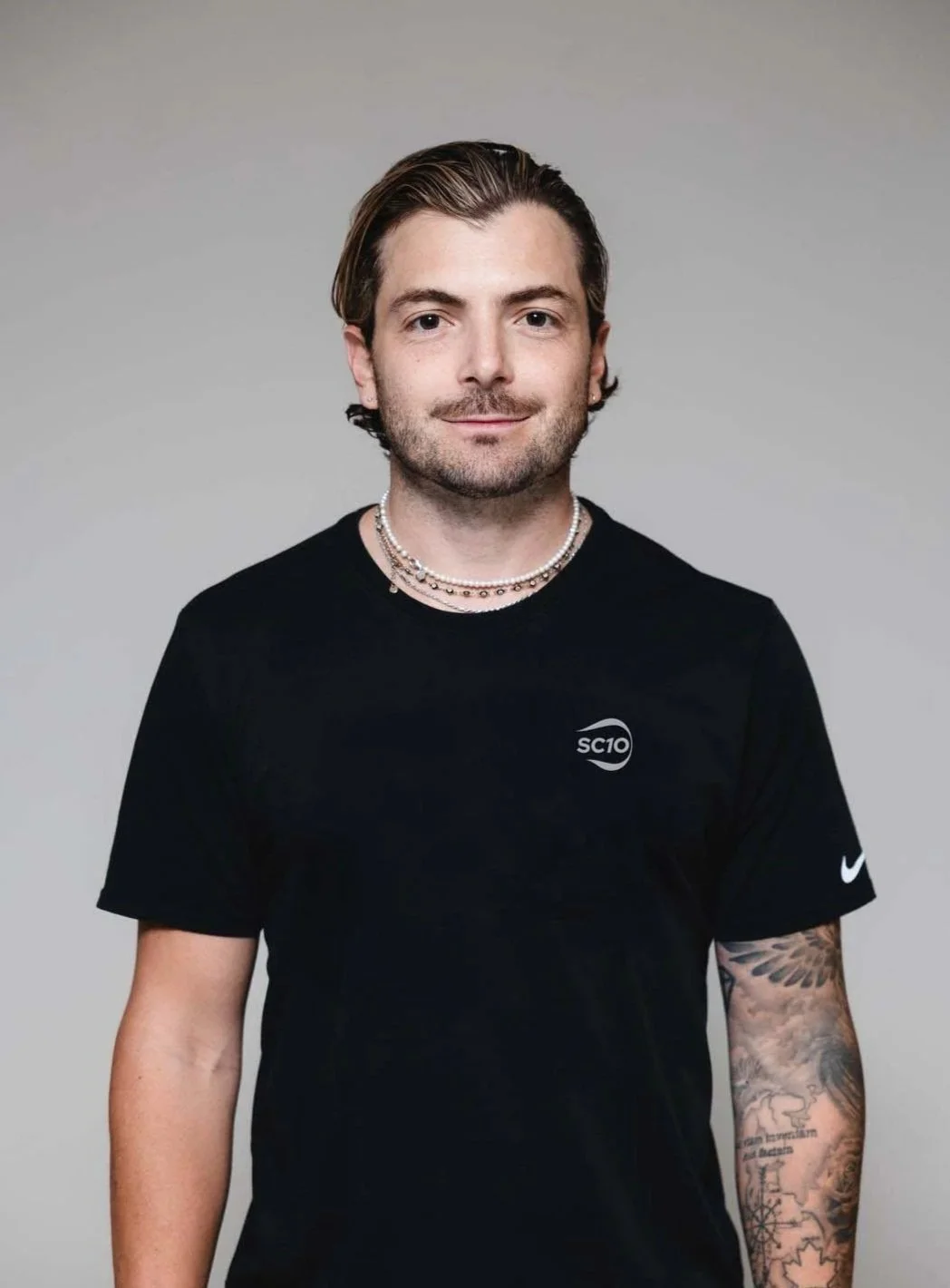 A young man with brown hair, beard, and mustache wearing a black T-shirt with 'SC10' logo and a Nike swoosh, accessorized with layered pearl necklaces, standing against a plain gray background.