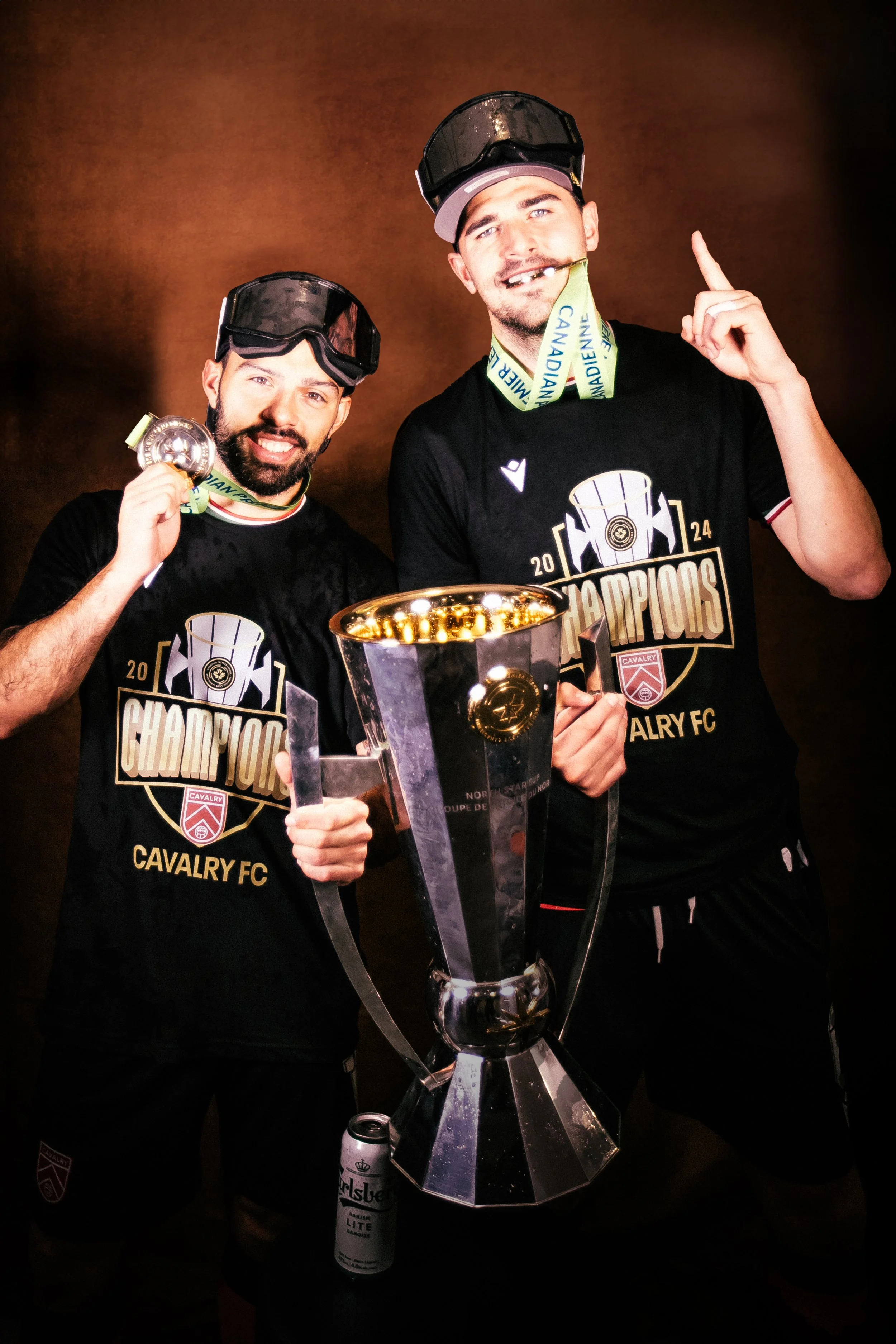 Sergio Camargo celebrating with a trophy, medals, and wearing black T-shirts that read 'Champions Cavalry FC.' They are wearing black hats with protective gear on top, smiling, and standing in front of a dark background.