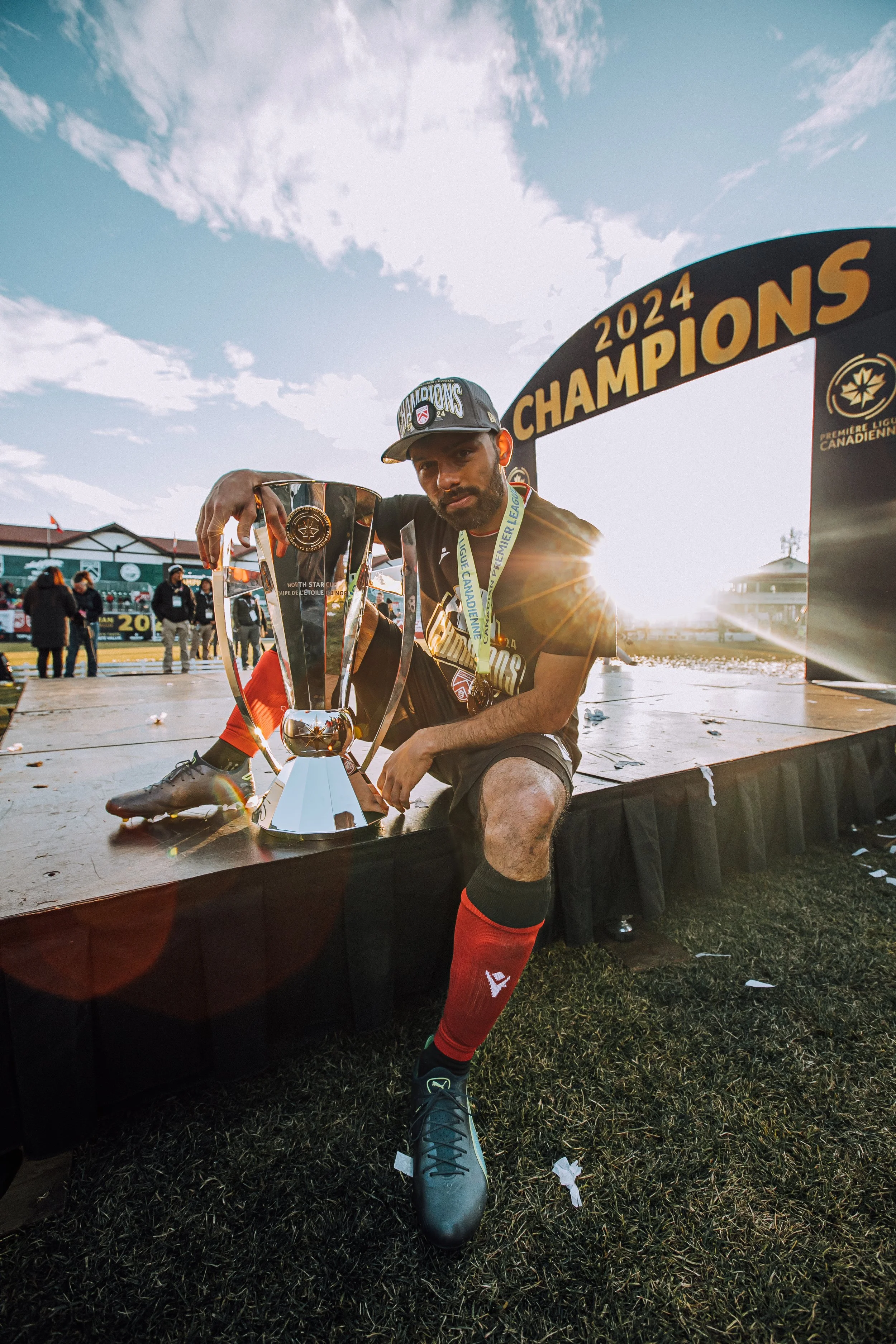 Sergio Camargo sitting on a stage, holding a trophy, with a medal around his neck. The backdrop sign reads '2024 Champions' and the logo of the Canadian Premier League. 