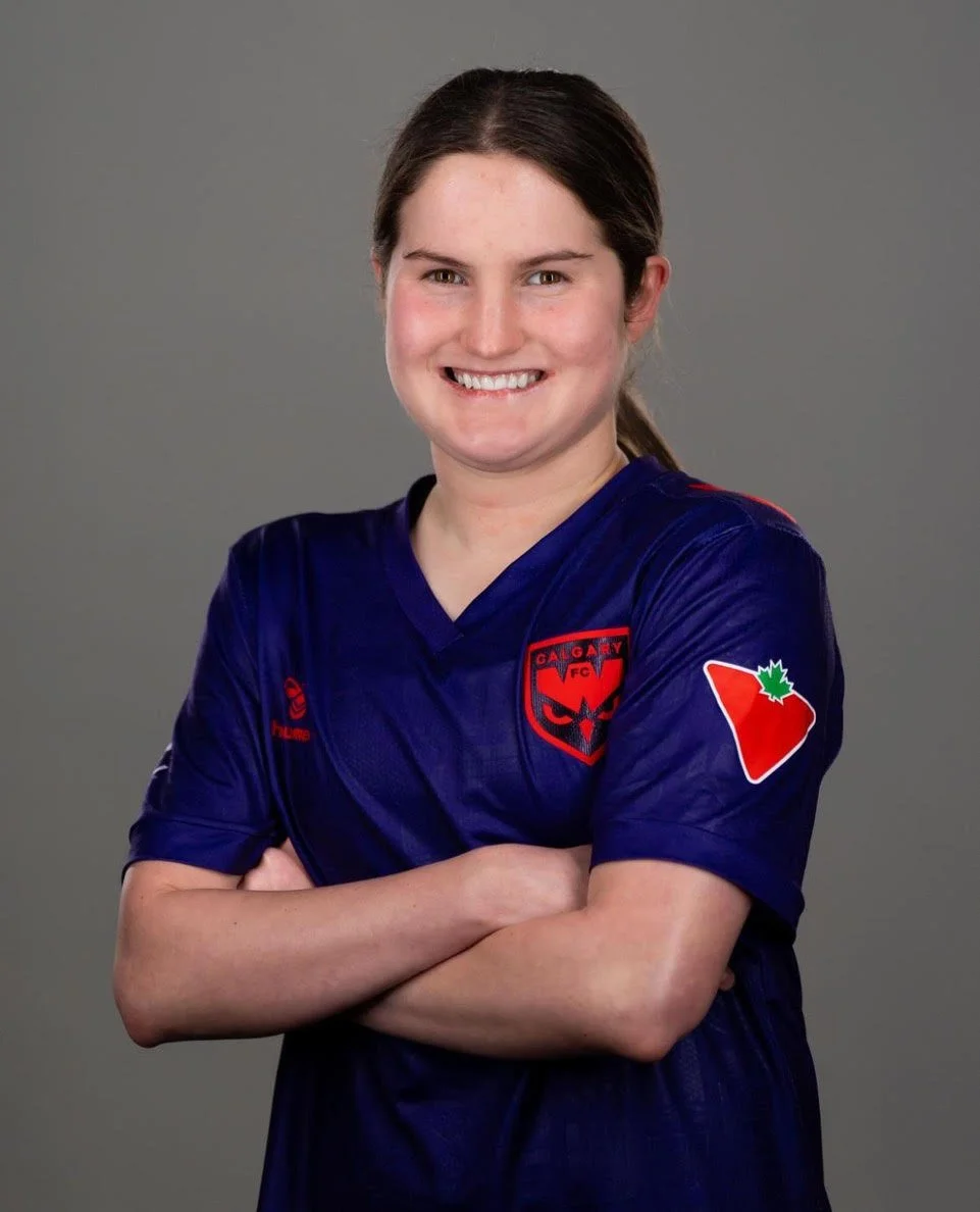Keelyn Stewart with dark hair smiling and arms crossed, wearing a blue sports jersey with a Calgary Wild FC logo and an Ontario patch on the sleeve, standing against a gray background.