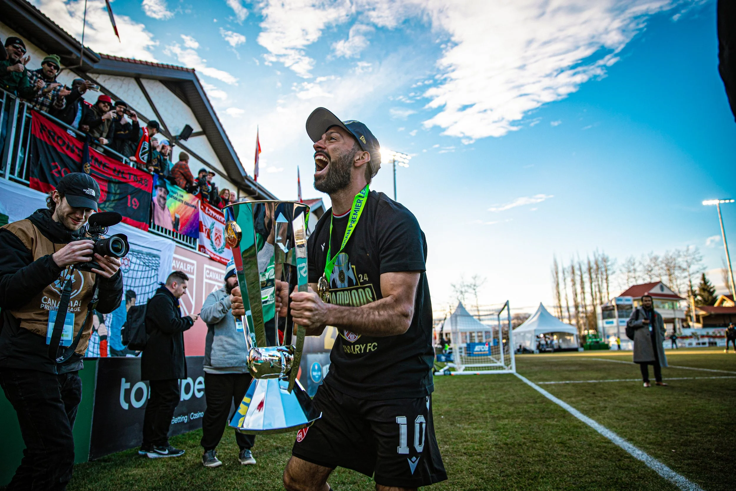 Sergio Camargo, a soccer player celebrating with a large trophy after a game, while a photographer captures the moment and spectators cheer in the background.