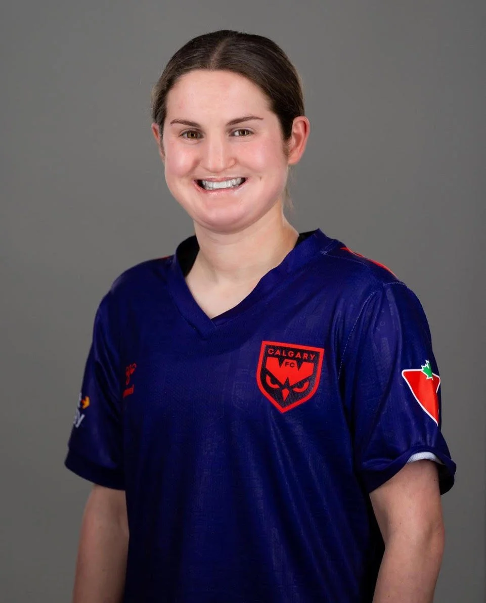 Taegan Stewart smiling woman with brown hair tied back, wearing a navy blue sports jersey with the Calgary Wild FC logo on it, standing against a gray background.