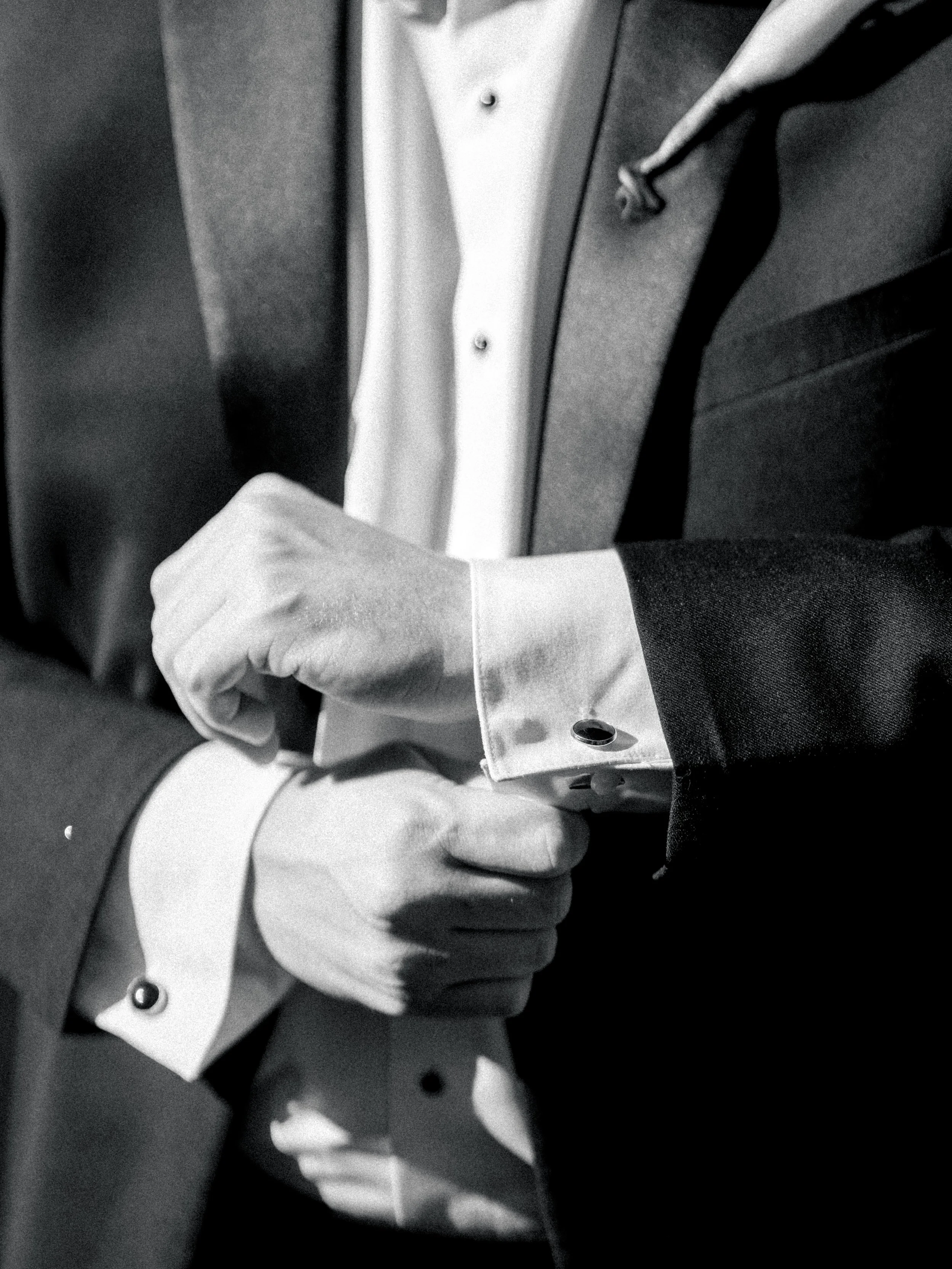 Close-up of a man adjusting his cufflinks, dressed in a tuxedo with a white shirt and black jacket, black tie, in black and white.