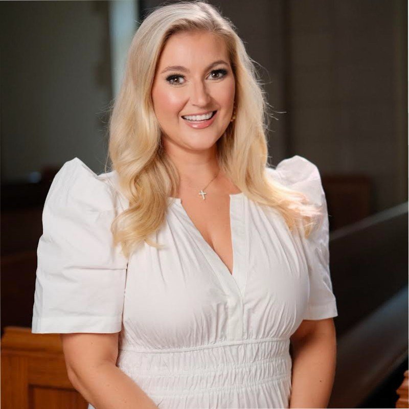 A smiling blonde woman with wavy hair wearing a white dress and a cross necklace.